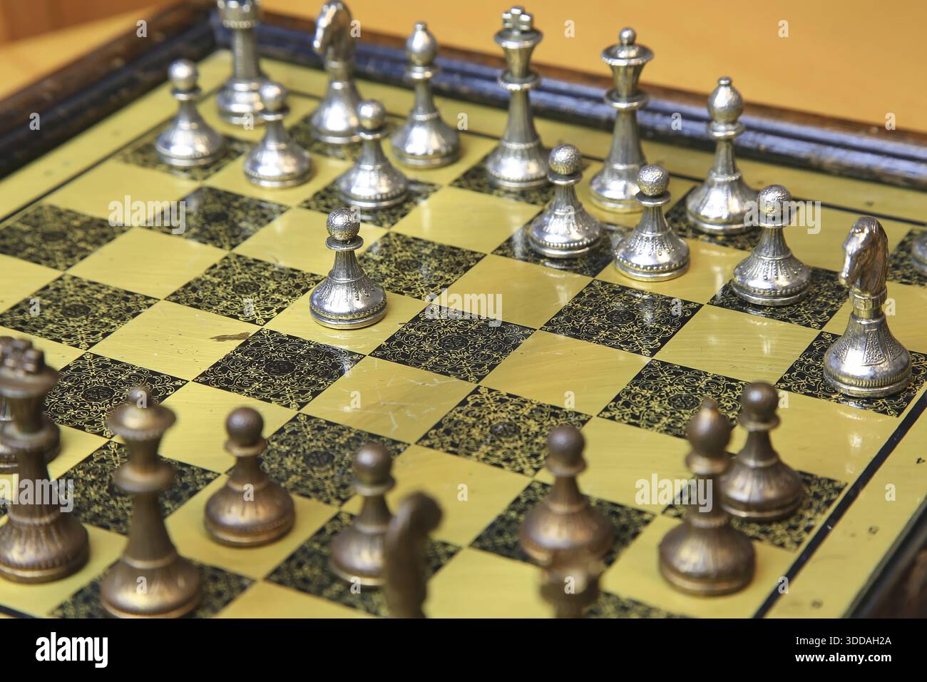 Ein feines Schachspiel auf einer gelben Pinnwand zieht die Aufmerksamkeit auf sich. Metallische Figuren in Silber und Bronze befinden sich in einer perfekten Ausgangsposition und reflektieren Stockfoto