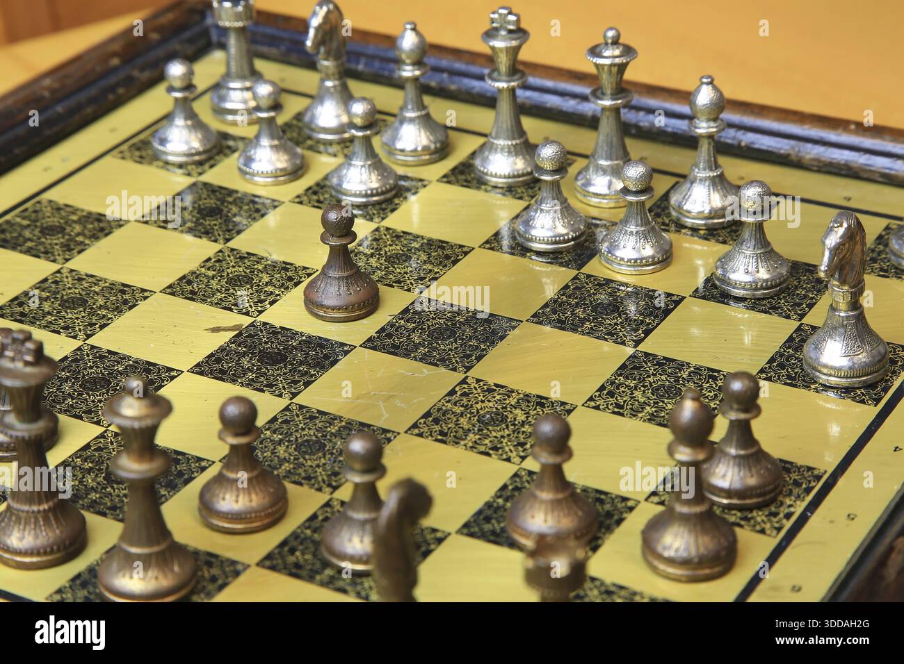 Ein feines Schachspiel auf einer gelben Pinnwand zieht die Aufmerksamkeit auf sich. Metallische Figuren in Silber und Bronze befinden sich in einer perfekten Ausgangsposition und reflektieren Stockfoto