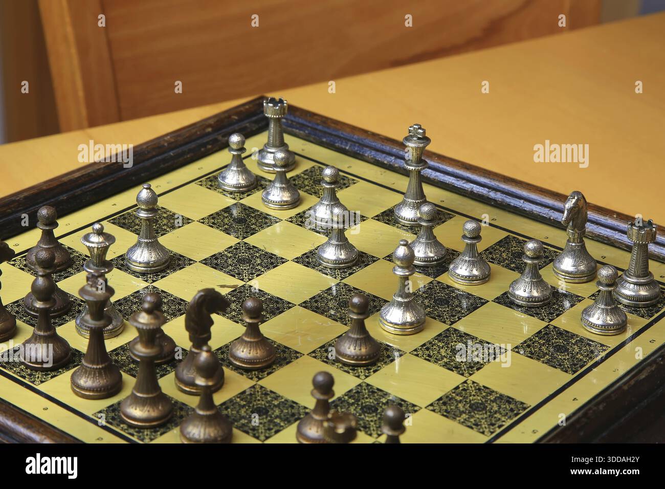 Ein feines Schachspiel auf einer gelben Pinnwand zieht die Aufmerksamkeit auf sich. Metallische Figuren in Silber und Bronze befinden sich in einer perfekten Ausgangsposition und reflektieren Stockfoto