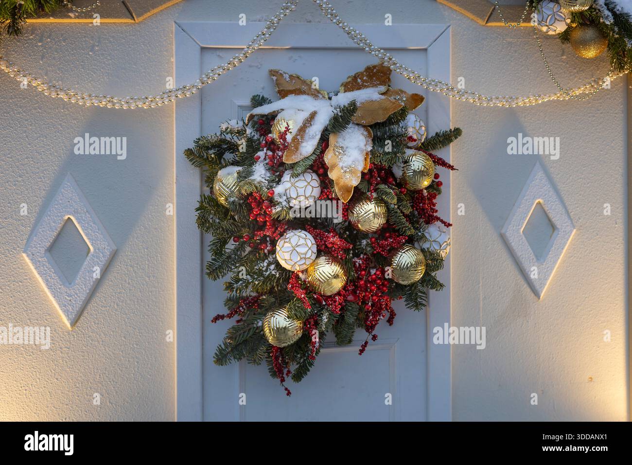 Ein Weihnachtskranz mit goldenen und weißen Ornamenten, roten Beeren und satinierten Blättern hängt an einer weißen Tür mit diamantförmigen Paneelen. Stockfoto