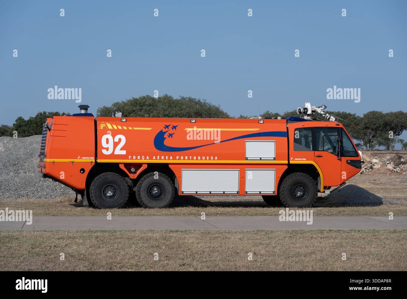 Beja, Portugal - 18. Juni 23: Ein leuchtend orangefarbener Feuerwehrwagen mit markantem Logo, auf einem grasbewachsenen Gelände, mit Bäumen und einer Baustelle im Ba Stockfoto