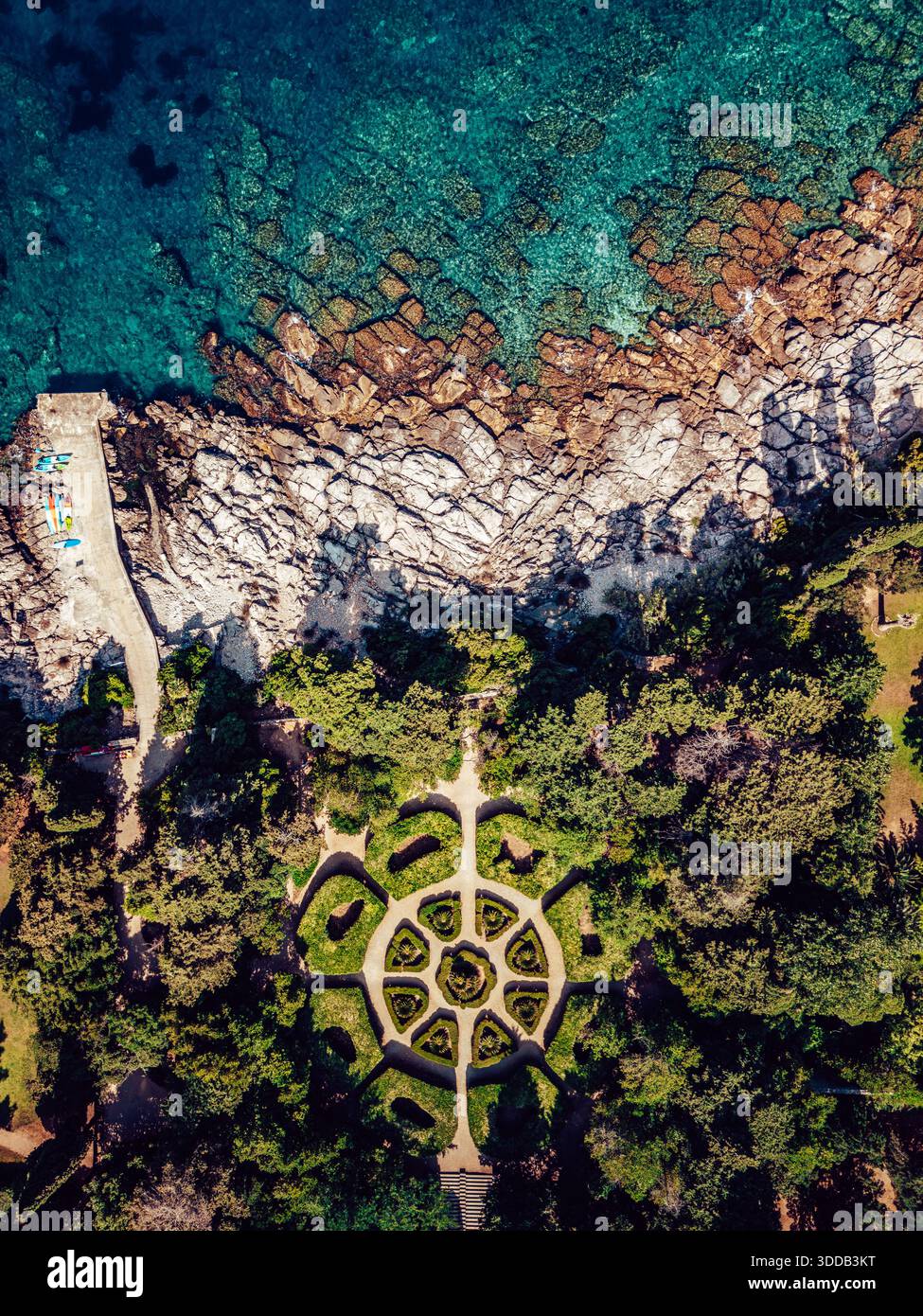 Blick aus der Vogelperspektive auf das türkisfarbene Meer, das die zerklüfteten, weißen Felsen küsst, während ein sorgfältig gestalteter Garten inmitten üppiger Vegetation in Dubrovnik, Dubrovnik liegt Stockfoto