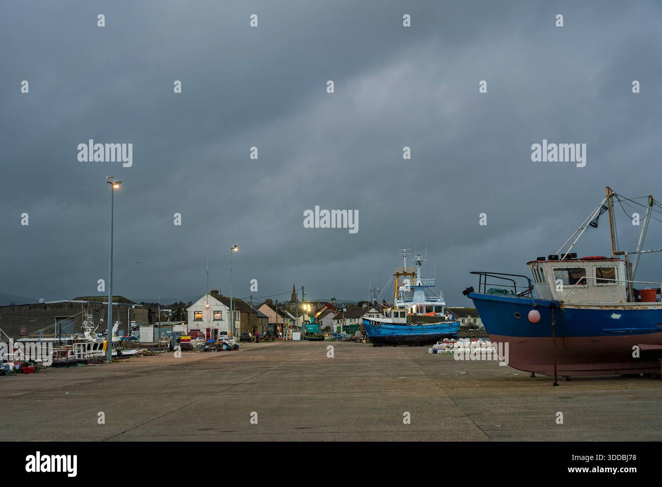 In der Abenddämmerung stehen auf dem Arklow-Festplatz funktionierende Schleppnetzfischer mit Stadtlichtern und schwerem grauem Himmel über dem Hafen. Stockfoto