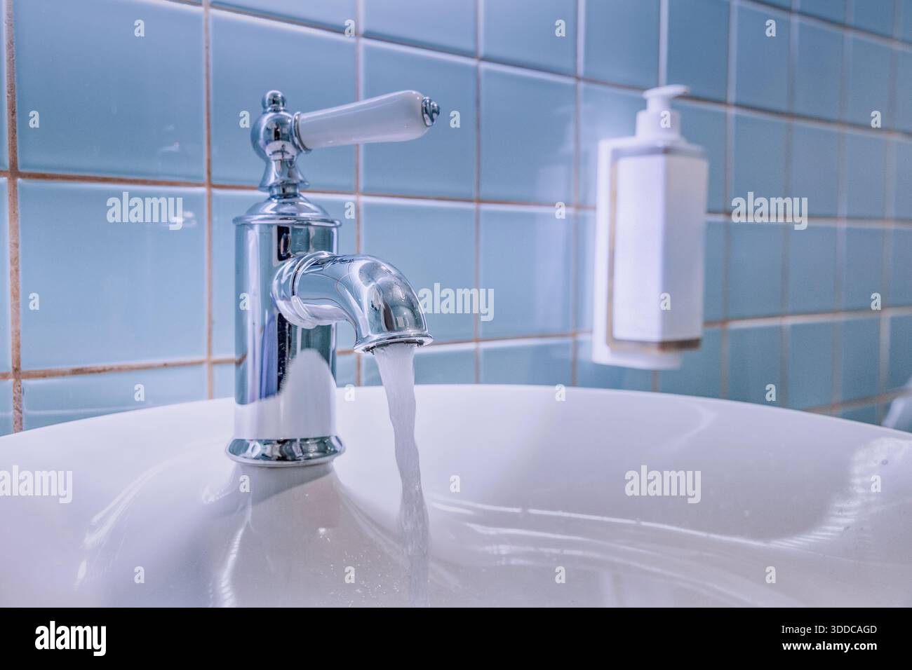 Fließendes Leitungswasser strömt in ein sauberes weißes Keramikbecken mit einem Vintage-Chromhahn gegen die blau geflieste Badezimmerwand Stockfoto
