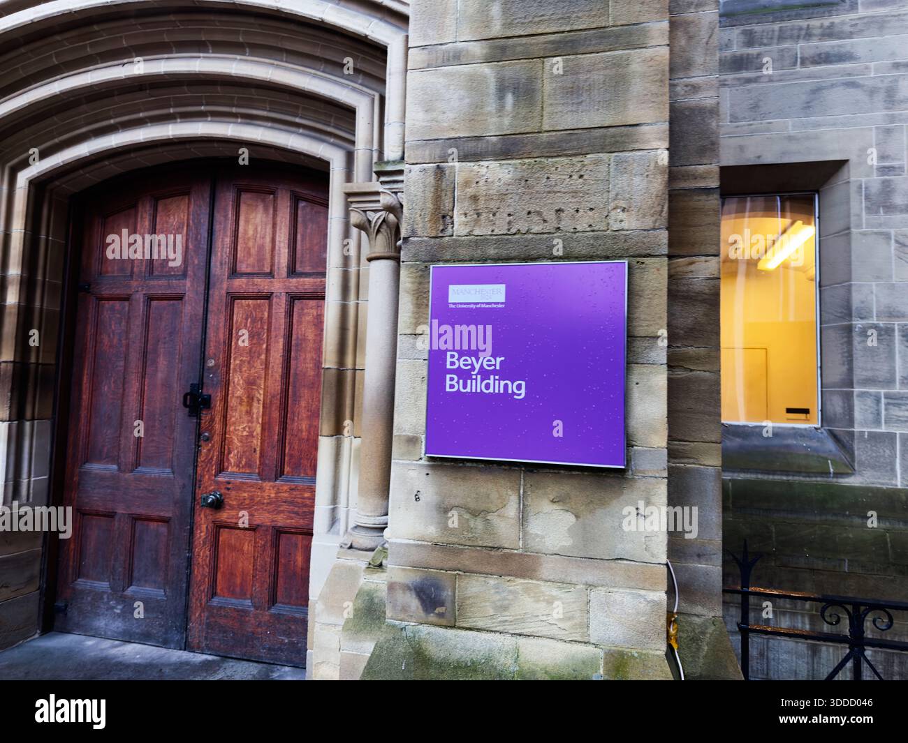 Manchester, Großbritannien - 06. Dezember 2025: Steingebäude in Manchester mit einer bogenförmigen Holztür und einem violetten Charles Beyer Building Schild, urbaner Campus-Atmosphäre. Stockfoto