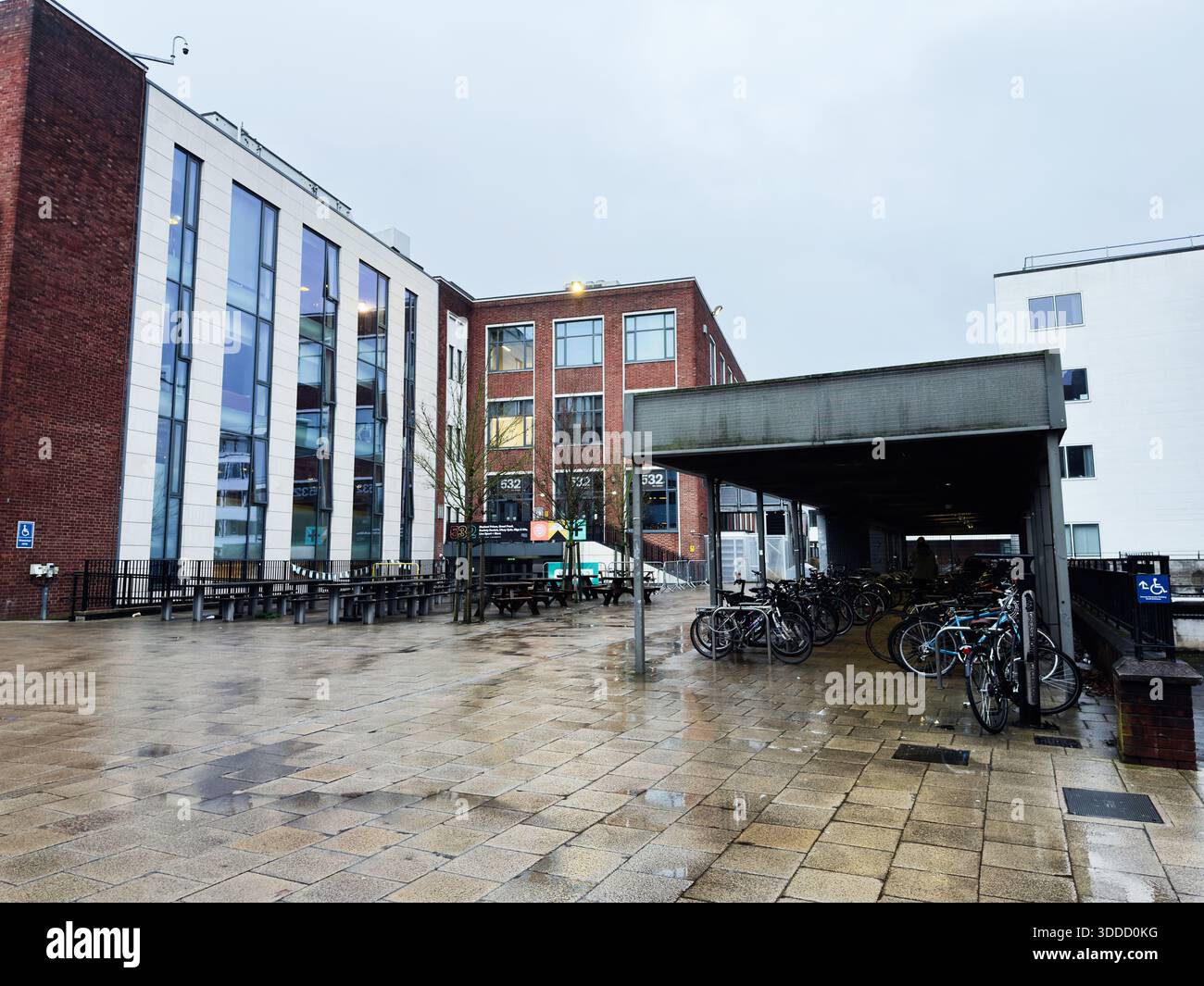 Manchester, Großbritannien - 06. Dezember 2025: Modernes Universitätsgebäude aus Backstein und Glas mit überdachtem Fahrradbereich und nassem Gehweg. Stockfoto