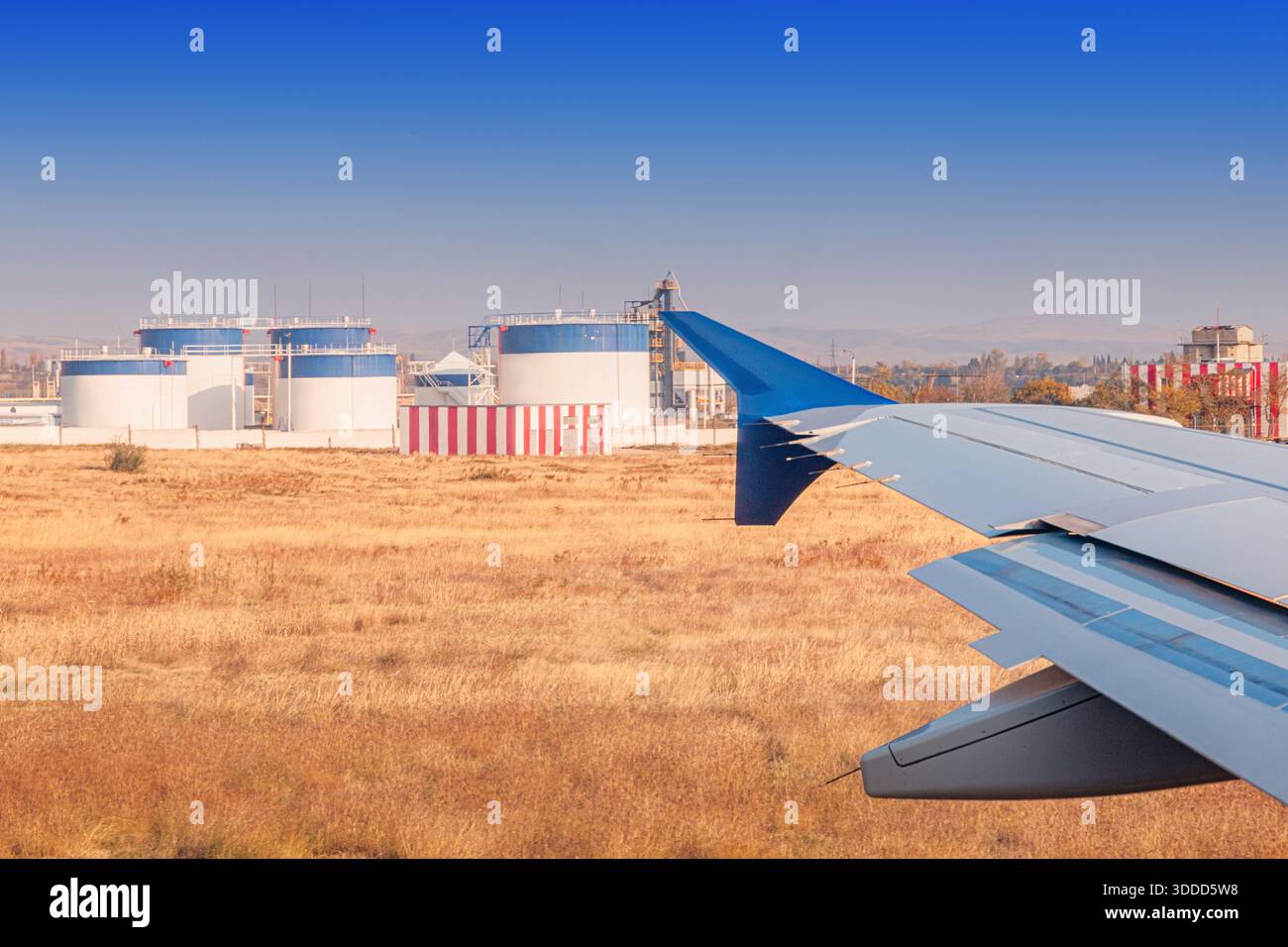 Flugzeugflügel und Flugzeugklappen sind sichtbar, wenn sie sich großen Treibstofftanks und Industrieanlagen neben einem Flughafen nähern Stockfoto