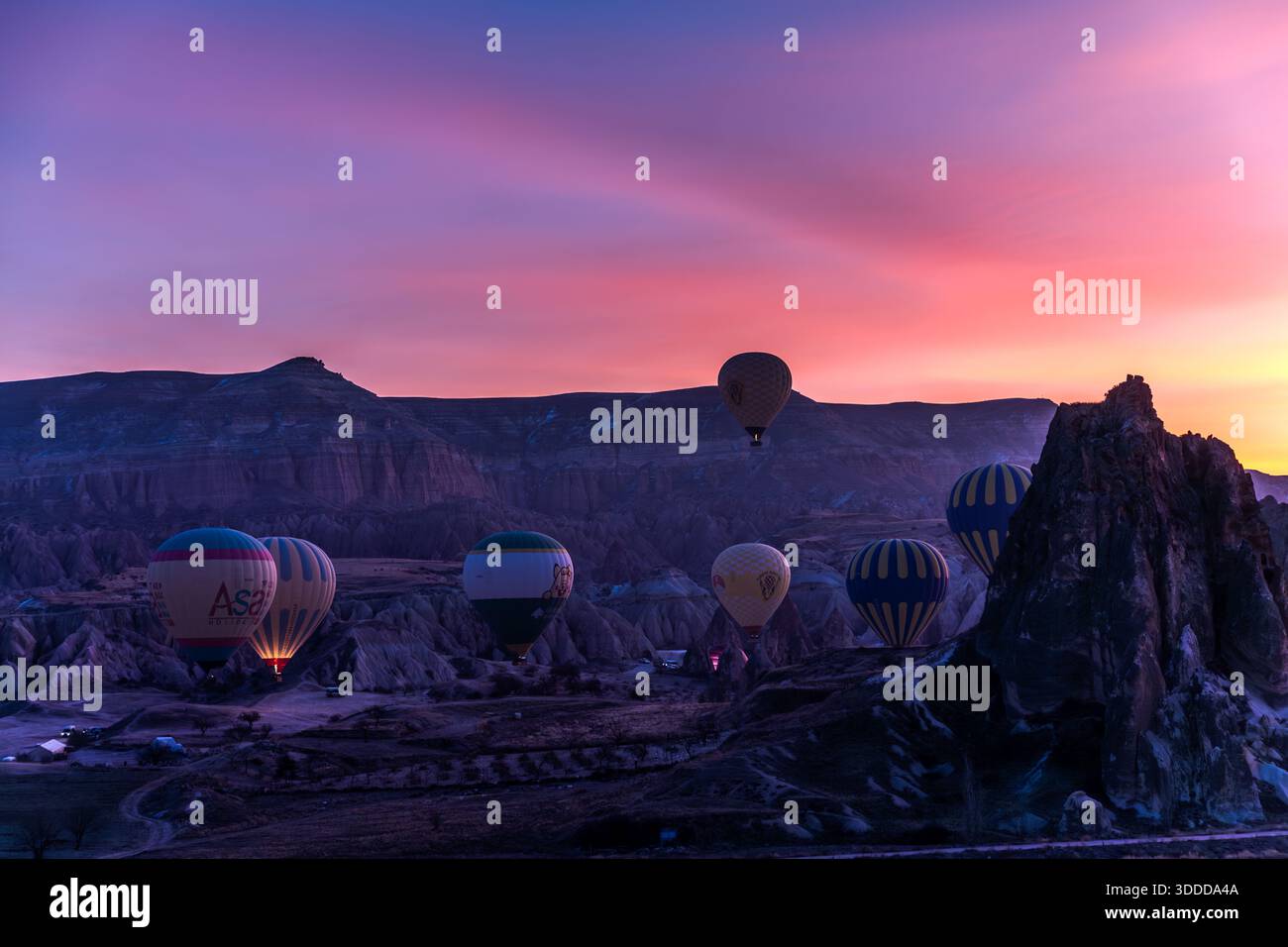 Heißluftballone steigen bei Sonnenaufgang über den Felsformationen und Feenschornen von Göreme, Kappadokien, Türkei; das Bild zeigt die charakteristische Tuffsteinlandschaft der Region mit einem markanten Felsnadel. Kommerzielle Ballonflüge über Kappadokien datieren zurück auf 1991, gegründet von der britischen Pilotin Kaili Kidner und ihrem schwedischen Ehemann Lars-Eric Möre; heute betreiben etwa 27 lizenzierte Unternehmen rund 250 Ballone mit bis zu 156 Flügen und 3.000 bis 4.000 Passagieren pro Tag. Stockfoto