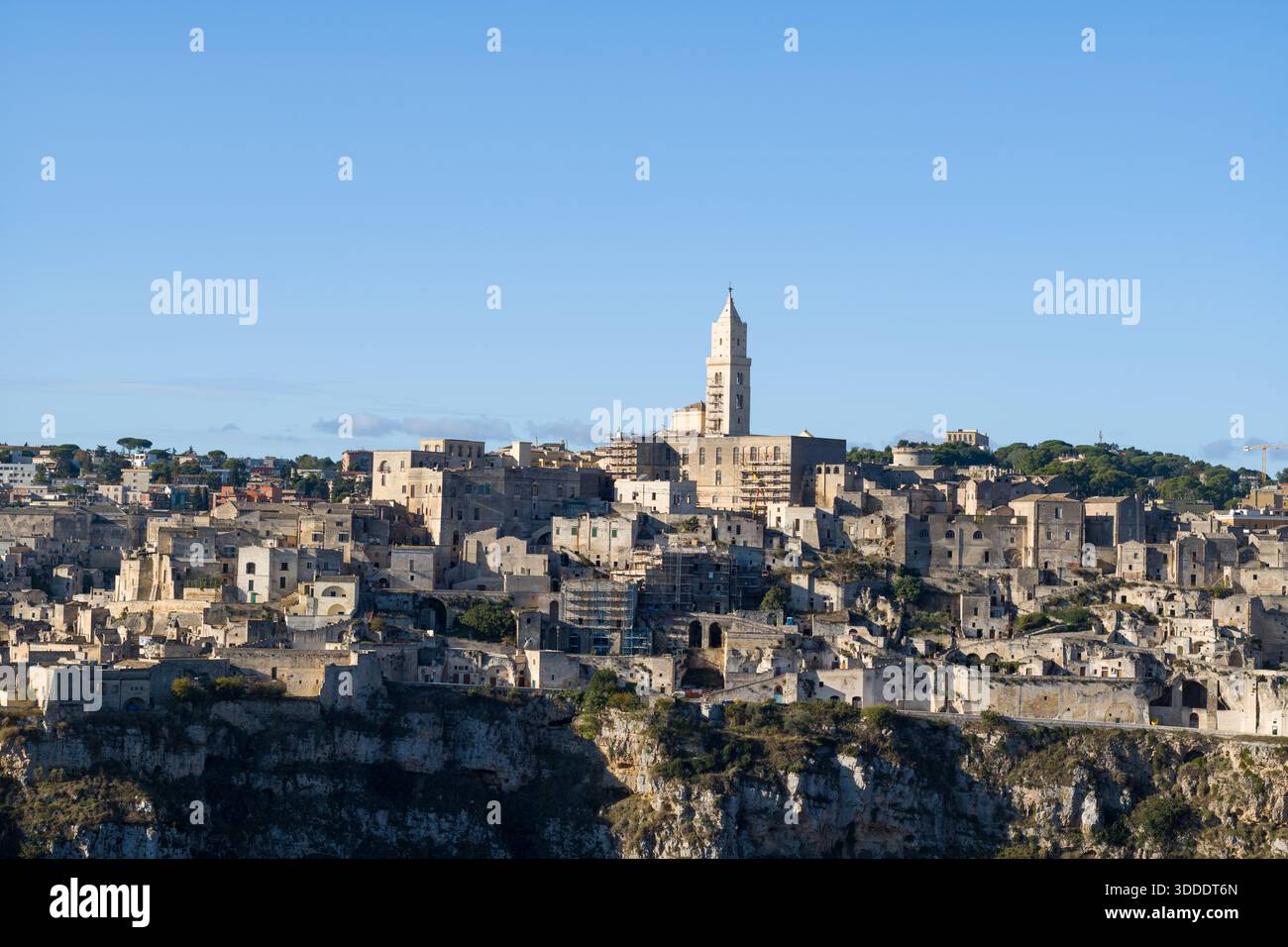 Historische Steinbauten von Matera erheben sich über zerklüftete Klippen, wobei der markante Turm der Kathedrale vor einem klaren blauen Himmel steht und das Sonnenlicht die antike Architektur hervorhebt. Stockfoto