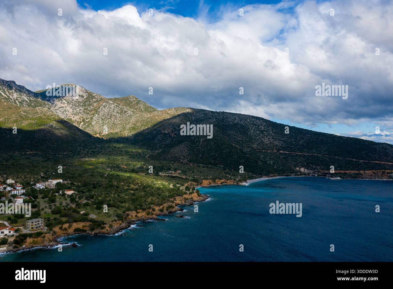 Eine zerklüftete Küste mit tiefblauem Wasser kurvt unter steilen, felsigen Bergen, die von Grün bedeckt sind. Sonnenlicht und Schatten spielen in der Landschaft unter einem Himmel voller strukturierter Wolken. Stockfoto