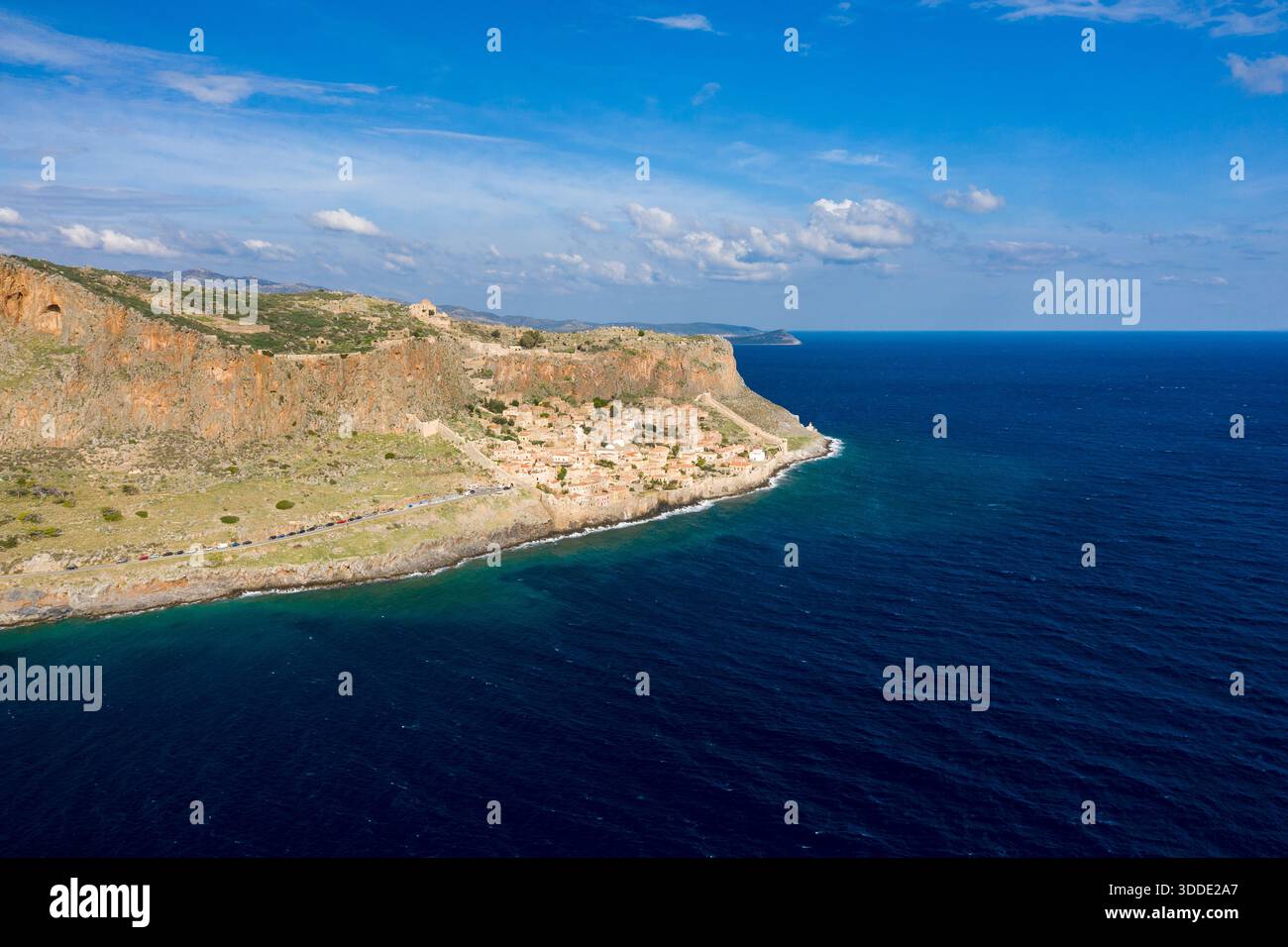 Blick aus der Vogelperspektive auf das historische Dorf Monemvasia, eingebettet unter steilen orangen Klippen am leuchtend blauen Mittelmeer. Die Szene bietet zerklüftete Felsstrukturen, klares Wasser und einen hellen Himmel mit verstreuten Wolken. Stockfoto