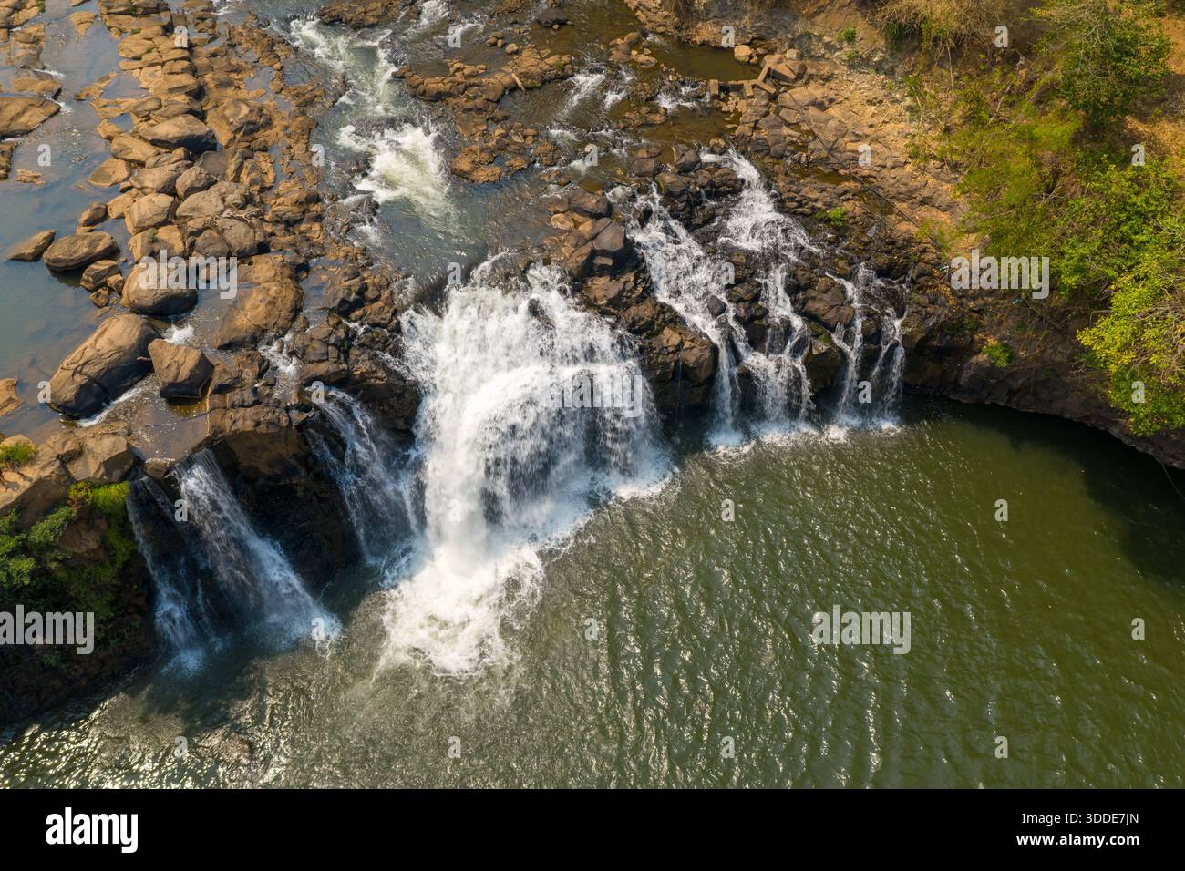 Die atemberaubende Perspektive des Wasserfalls Tad Lo, während das Wasser über zerklüftete Felsen in ein breites Becken im Süden von Laos stürzt. Sonnendurchflutete Texturen heben den Kontrast zwischen fließendem Wasser, freiliegendem Stein und umliegendem Grün hervor. Stockfoto