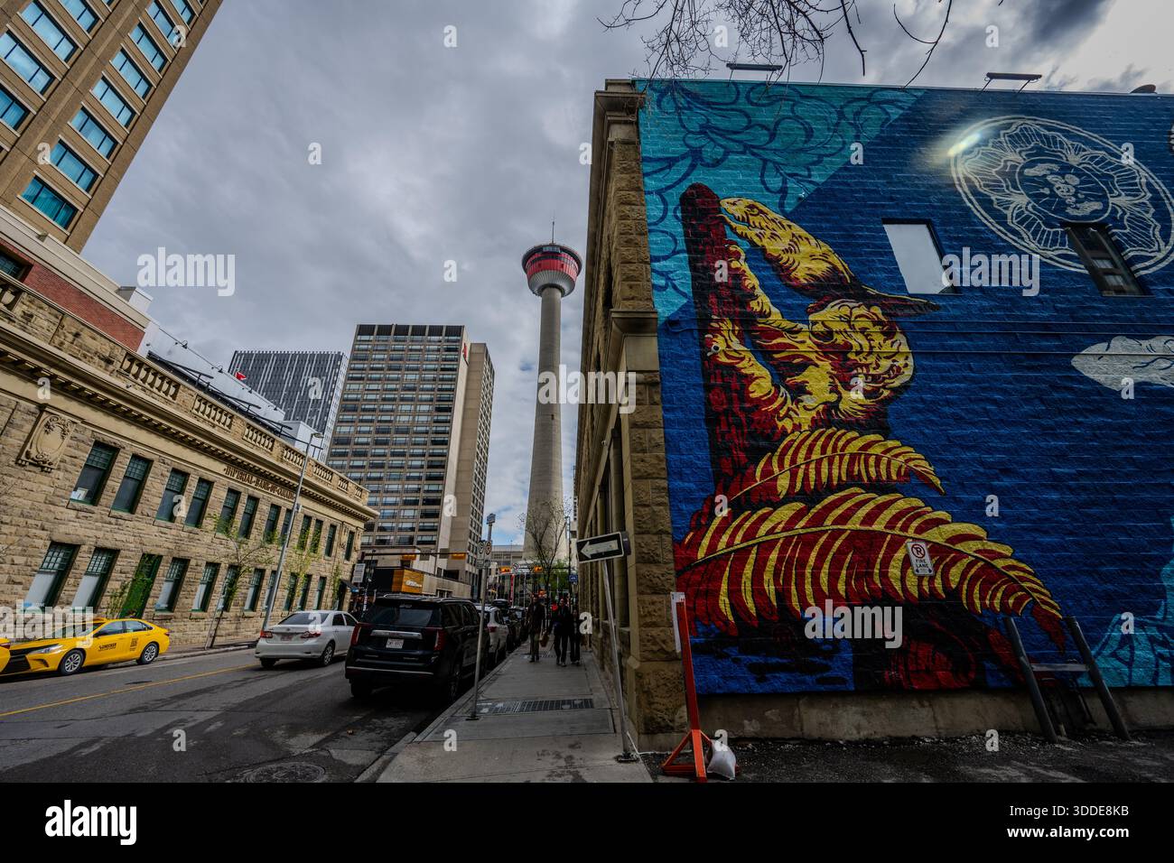 Ein farbenfrohes Wandgemälde bedeckt die Seite eines Gebäudes, während der Calgary Tower sich zwischen den umliegenden Bürogebäuden erhebt und parkende Autos die Straße entlang der Innenstadt säumen. Stockfoto