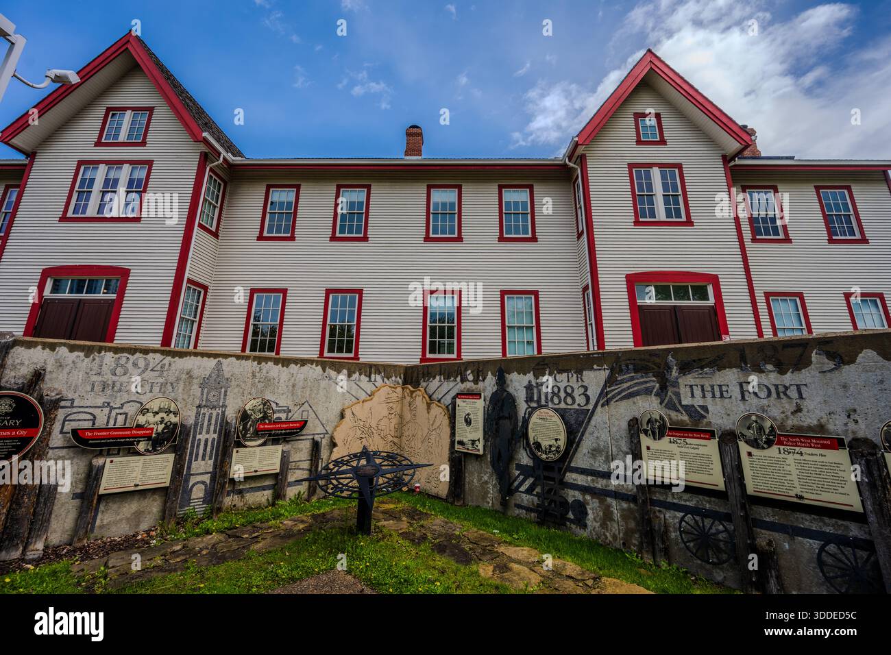 Ein großes weißes Schalengebäude mit roten Zierleisten steht hinter einer Betonwand, die mit historischen Wandmalereien und interpretierenden Tafeln auf einem grasbewachsenen Weg i bedeckt ist Stockfoto