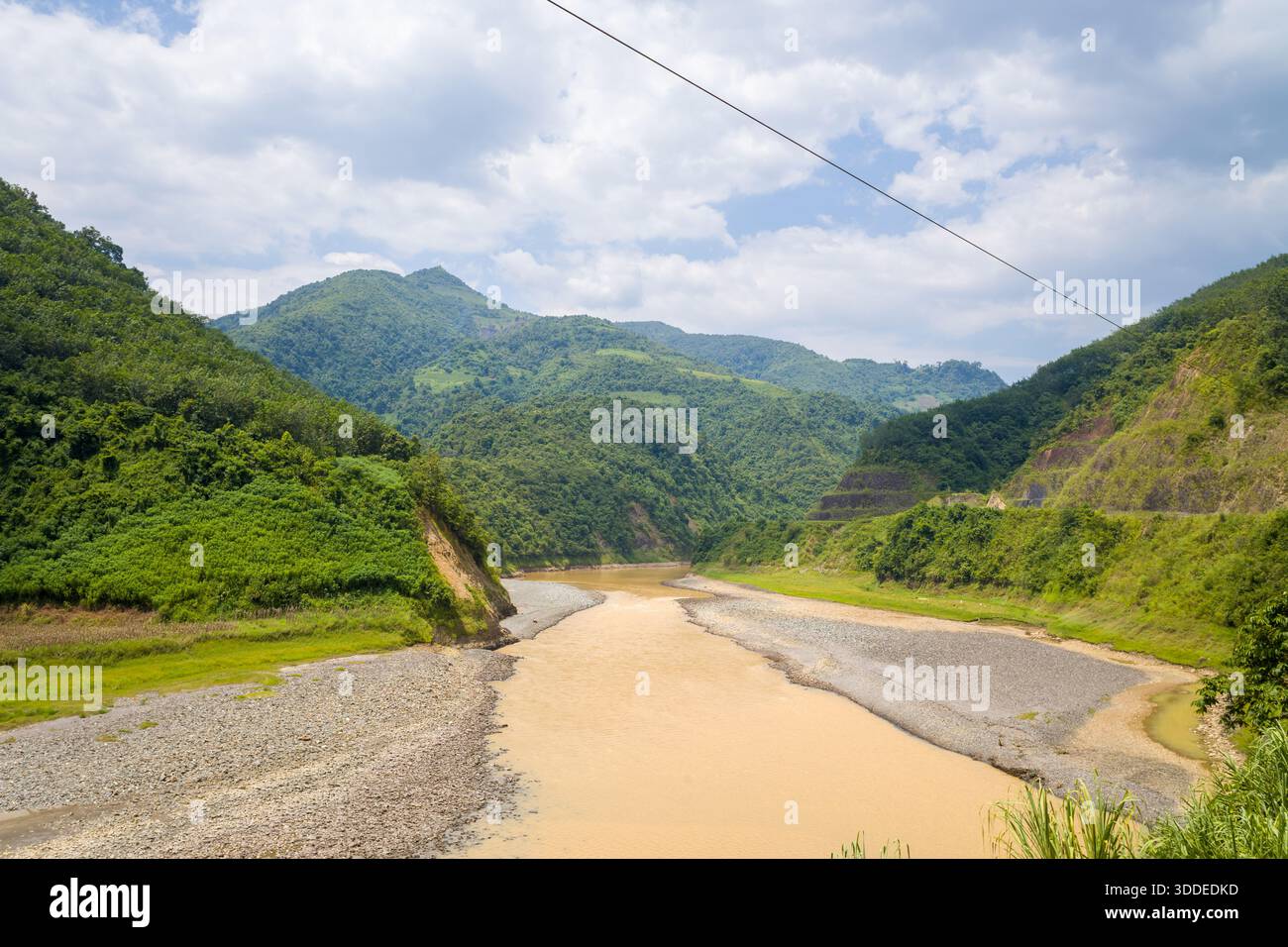 Ein schlammiger Fluss fließt durch ein weites Tal, das von üppigen grünen Bergen im Norden Vietnams flankiert wird. Helles Tageslicht und verstreute Wolken unterstreichen die natürlichen Strukturen der Landschaft. Stockfoto