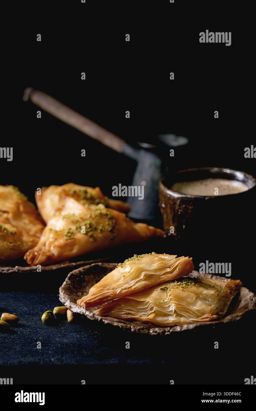 Hausgemachtes türkisches traditionelles Dessert Baklava mit Pistazien serviert auf Keramikplatte mit Nüssen, Tasse Kaffee, Vintage-Cezva auf schwarzem Holztisch. Kopierbereich Stockfoto