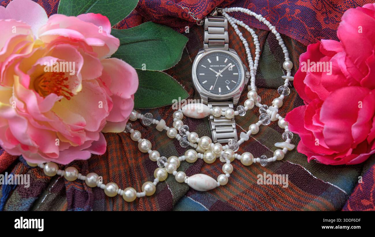 Armbanduhr aus Edelstahl, umgeben von Perlenketten und rosafarbenen Blumen auf gemustertem Stoff. Stockfoto