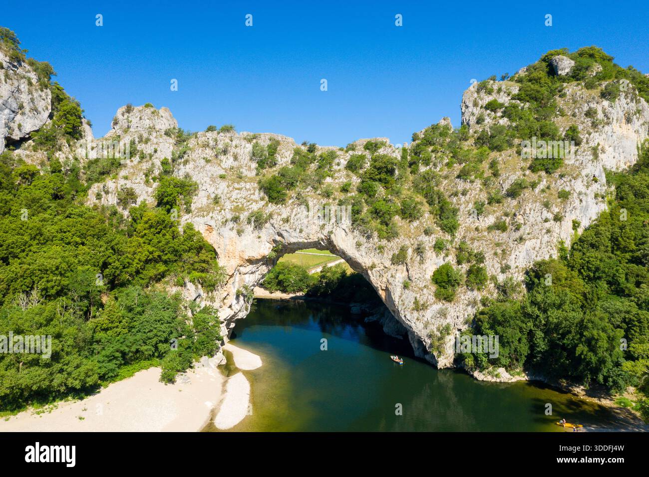 Ein weitläufiger natürlicher Kalksteinbogen erhebt sich über einem sanft geschwungenen Fluss, umgeben von dichtem grünen Wald und zerklüfteten Klippen unter einem klaren blauen Himmel in der Region Ardeche. Stockfoto