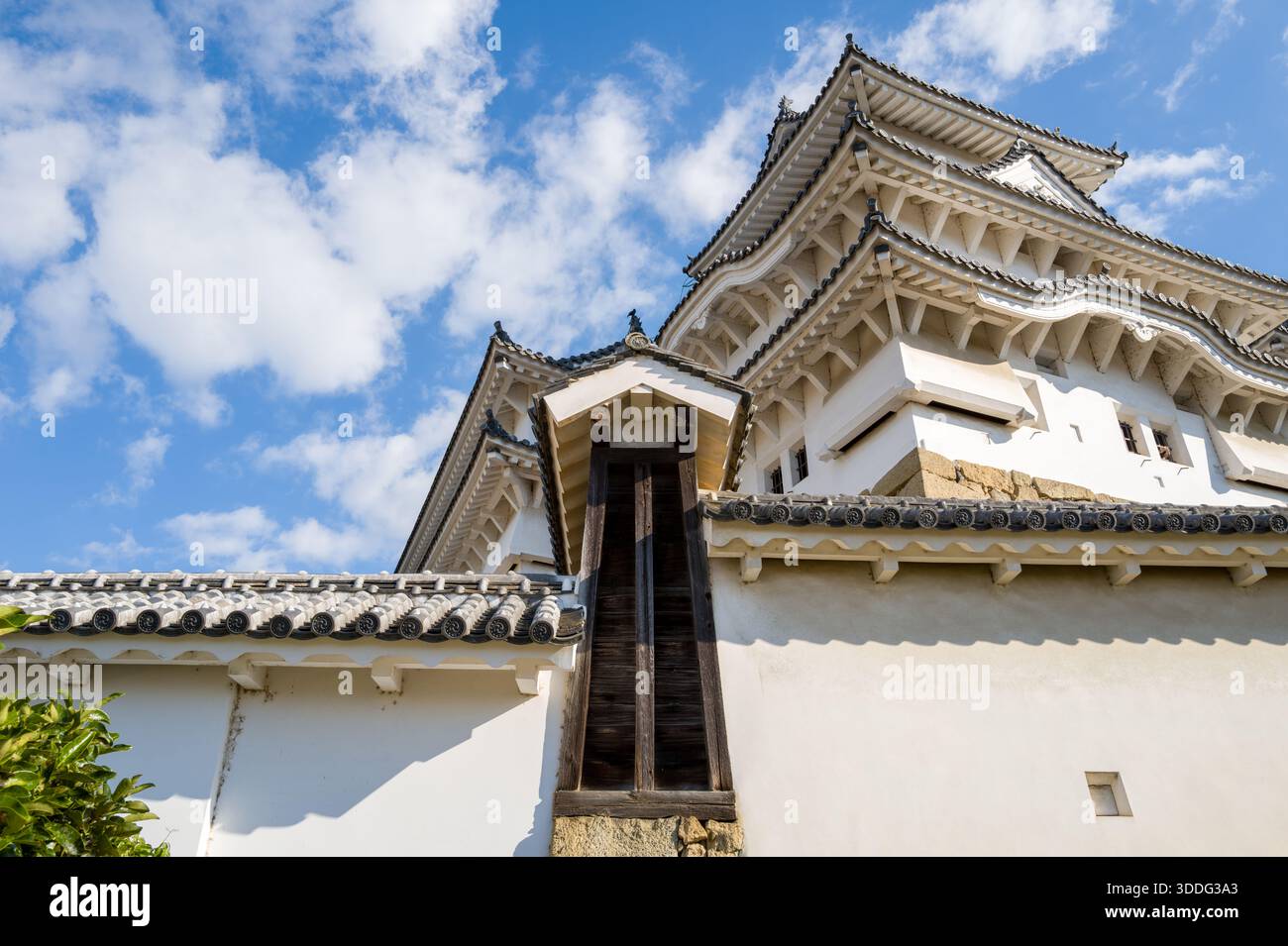 Beeindruckender Blick nach oben auf den weißen Turm der Burg Himeji mit mehrschichtigen Fliesentreffen und architektonischen Details aus Holz, der unter einem leuchtend blauen Himmel mit verstreuten Wolken liegt. Stockfoto