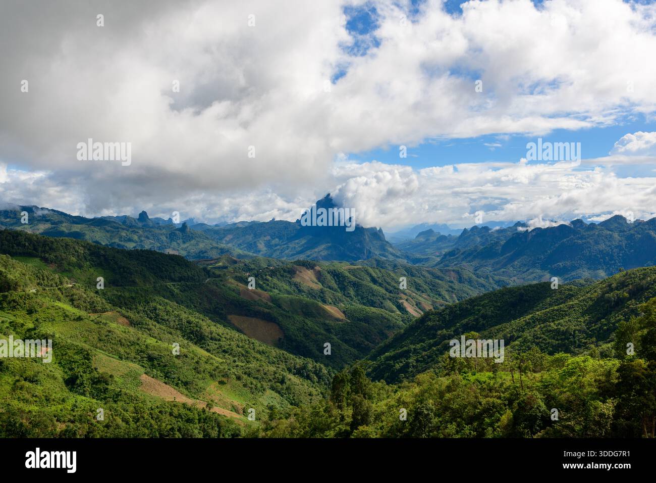 Sanfte grüne Hügel und dramatische Bergrücken erstrecken sich bis in die Ferne unter einem Himmel voller dicker Wolken und blauer Flecken. Die Landschaft bietet üppige Vegetation, tiefe Täler und einen markanten, zerklüfteten Gipfel, der teilweise in Nebel gehüllt ist. Stockfoto