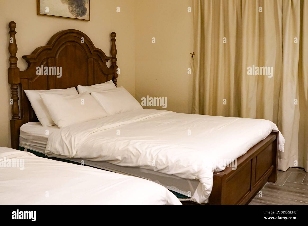 Schön gestaltetes Schlafzimmer mit zwei Betten, weißer Bettwäsche, hölzernem Kopfteil, beigefarbenen Vorhängen und abstrakten Wandbildern in einem warmen, einladenden Ambiente. Stockfoto