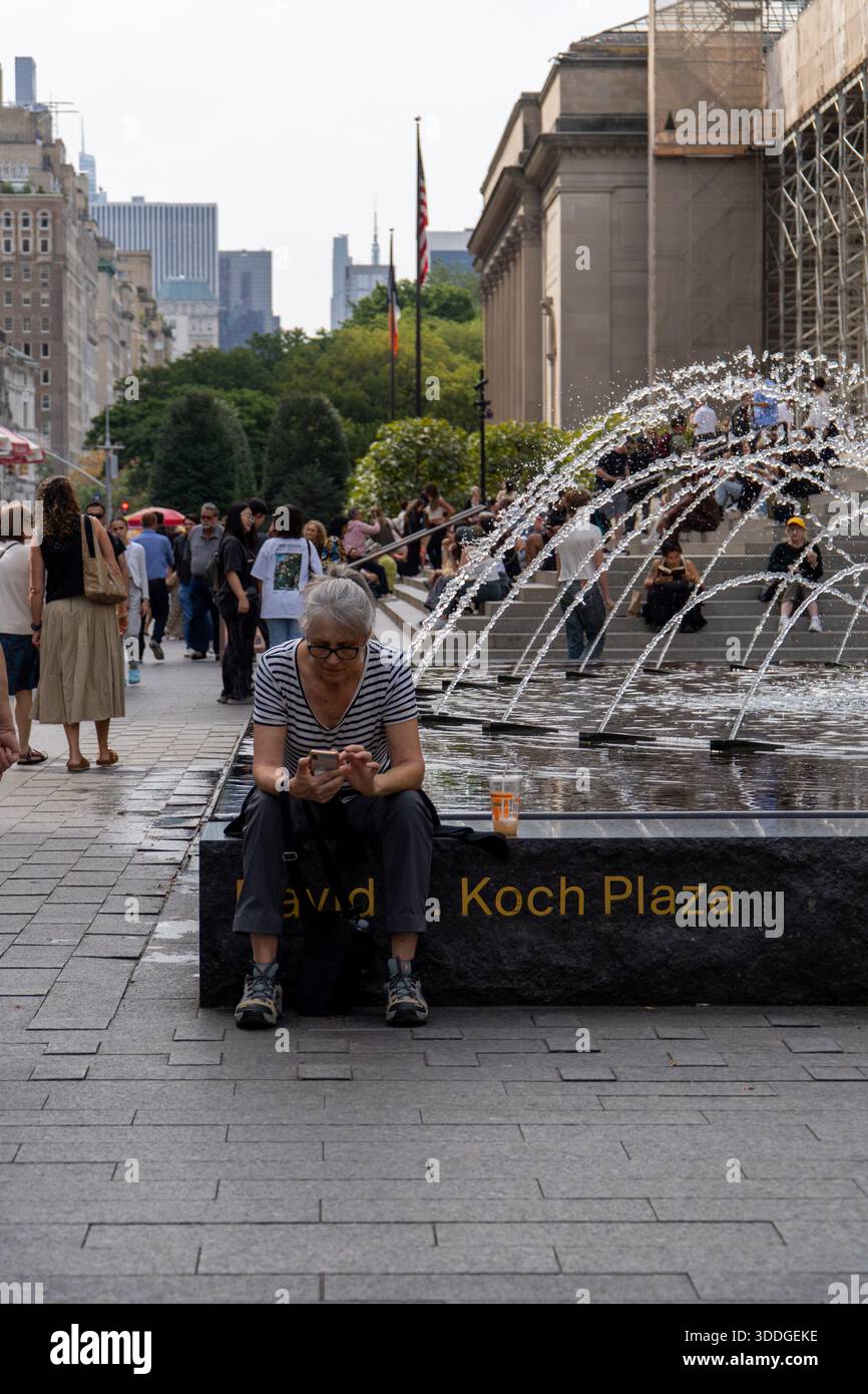 New York, USA. 30. September 2025. - Eine Frau sitzt am David H. Koch Plaza Brunnen, vertieft in ihr Smartphone, während die Leute sich entspannen und in der Nähe laufen. Stockfoto
