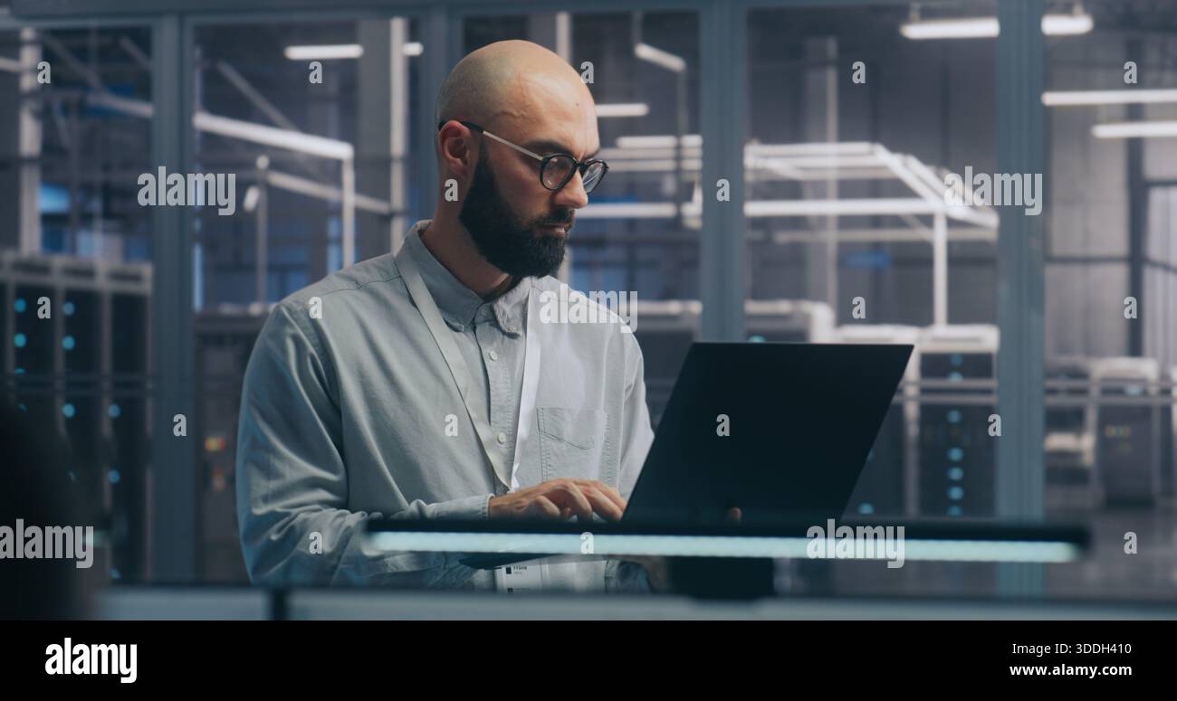 Fokussierter IT-Techniker mit Laptop in der Serverhalle. Mann in leichtem Shirt steht und arbeitet intensiv, Hintergrund zeigt Server-Racks modernes Rechenzentrum. Konzept vor-Ort-Diagnose und Systemwartung. Stockfoto