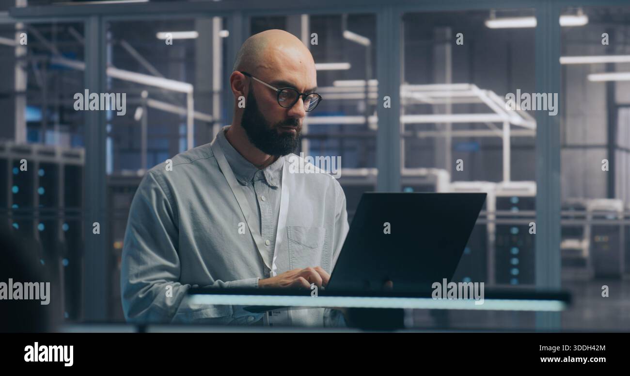 Fokussierter IT-Techniker mit Laptop in der Serverhalle. Mann in leichtem Shirt steht und arbeitet intensiv, Hintergrund zeigt Server-Racks modernes Rechenzentrum. Konzept vor-Ort-Diagnose und Systemwartung. Stockfoto