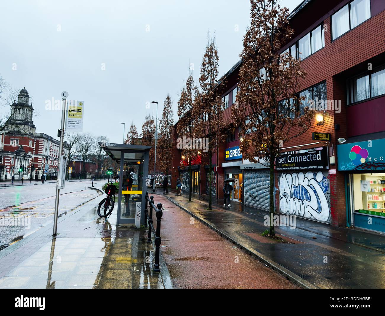Manchester, Großbritannien - 06. Dezember 2025: Regnerische Manchester Street mit Bushaltestelle, Radfahrern, Graffitis und Geschäften; städtisches Flair. Stockfoto