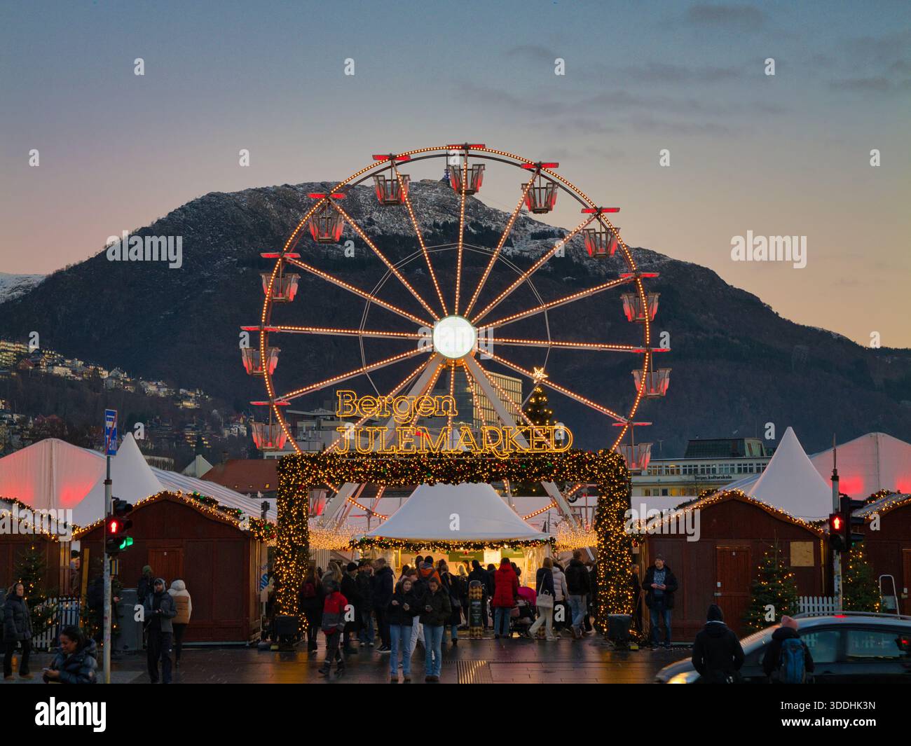 Bergen, Norwegen – 25. November 2025: In Bergen, Norwegen, betreten Menschenmassen den Julemark Christmas Market in Bergen, Norwegen, unter leuchtenden Lichtern und einem Riesenrad in der Abenddämmerung, mit Bergen im Hintergrund. Stockfoto