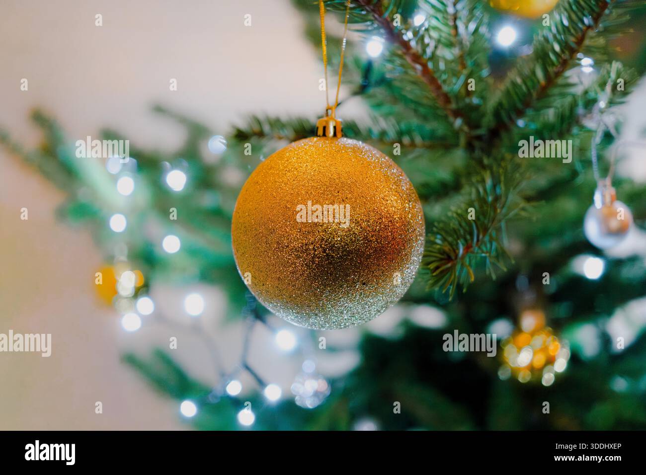Das Bild zeigt eine Nahaufnahme eines glitzernden goldenen Weihnachtsschmucks, der an einem geschmückten Weihnachtsbaum hängt. Stockfoto