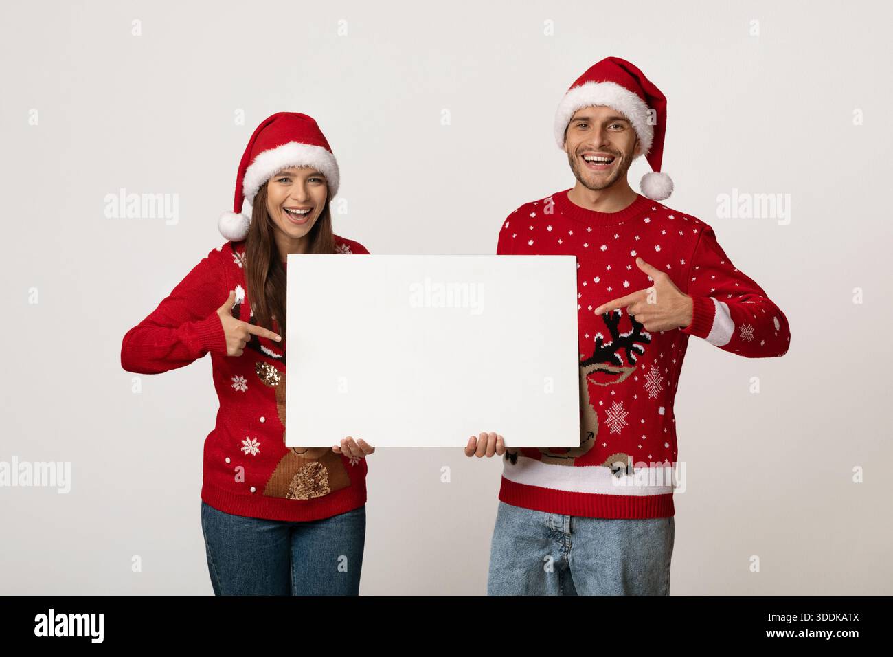 Glückliches Paar mit weihnachtlichen Pullover und Hüten, die ein leeres Schild für festliche Ankündigungen halten Stockfoto
