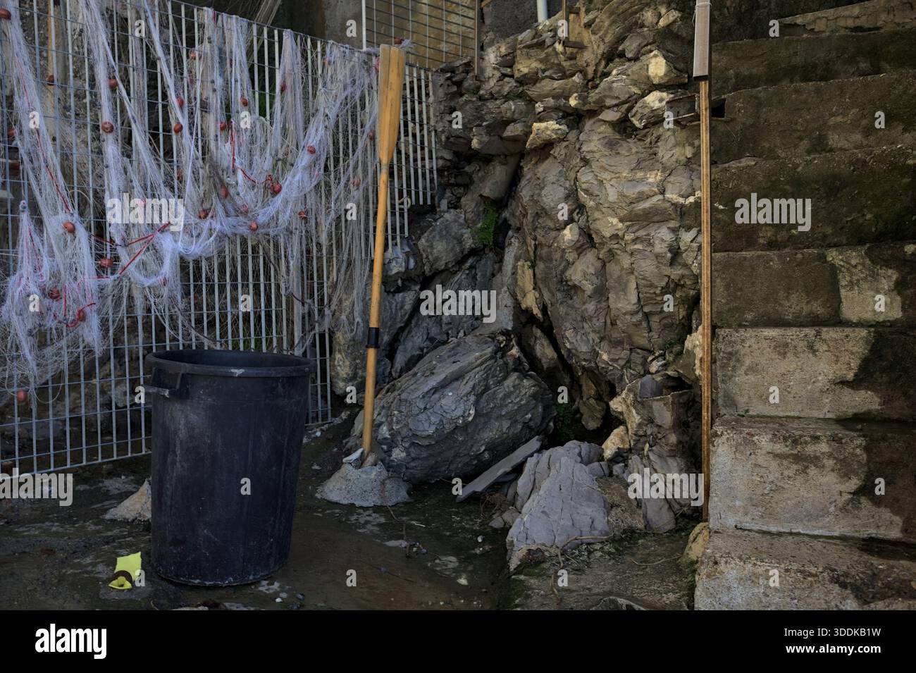 Schwarzer Mülleimer, ein stehendes Ruder und ein Fischernetz auf einem Zaun neben einer Treppe am Rand einer Steinmauer Stockfoto
