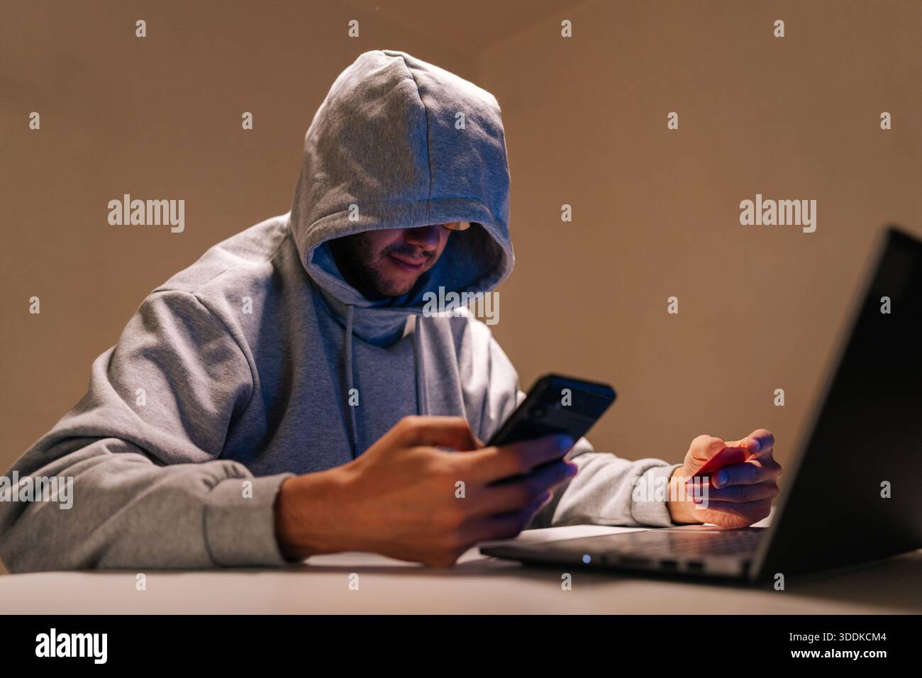 Eine Person mit Kapuze, die in Cyberkriminalität involviert ist, Online-Betrug und Datendiebstahl mit Mobiltelefon, Laptop und Kreditkarte in einer dunklen Umgebung durchführt Stockfoto