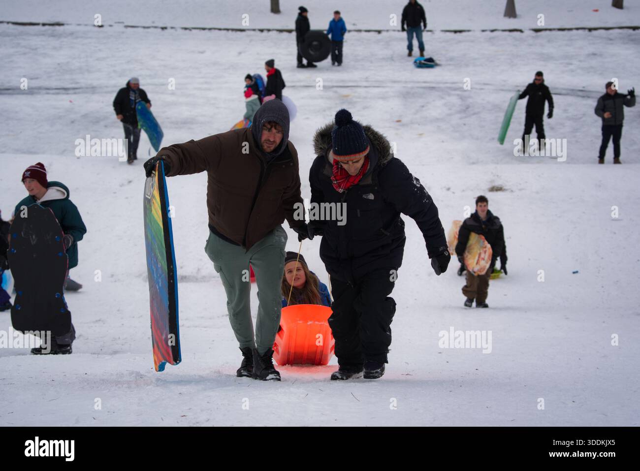 Die Leute fahren mit Schlitten und tragen Schlitten einen schneebedeckten Hügel hinunter. Ein Mann und eine Frau ziehen ein Kind in einem orangefarbenen Schlitten. Stockfoto