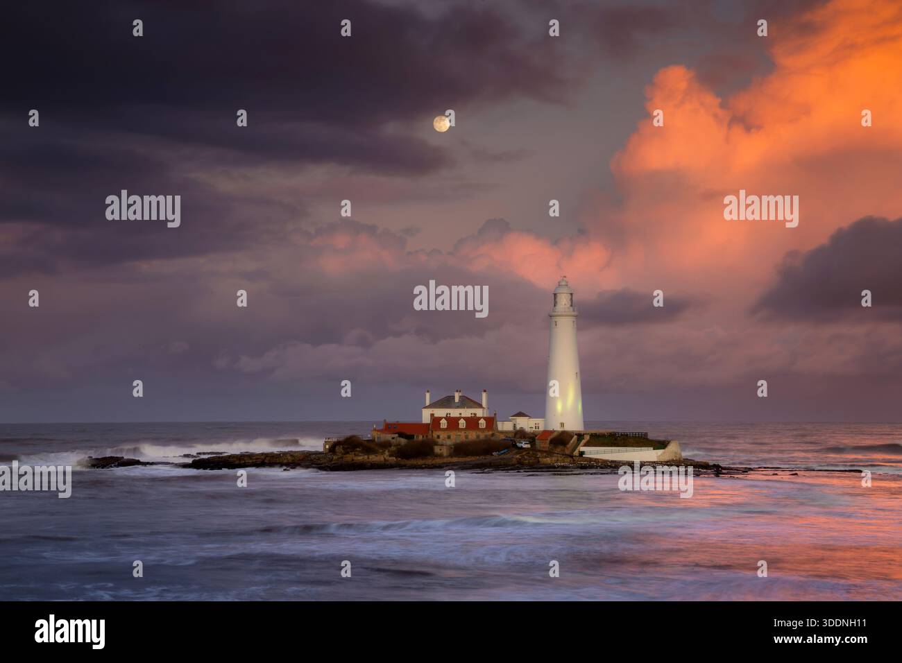 Ein einsamer Leuchtturm steht auf einer felsigen Insel, umgeben von einem Ozean mit stürzenden Wellen. Der Himmel zeigt atemberaubende Orange- und Violetttöne. Stockfoto