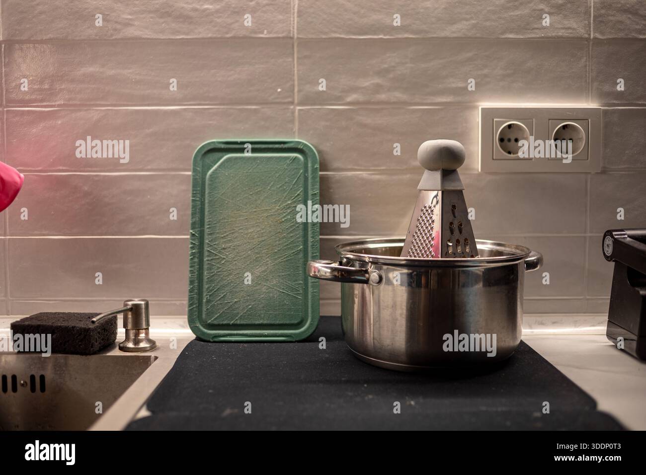 Küchenarbeitsplatte mit Edelstahltopf, Metallreibe innen, grünes Schneidebrett aus Kunststoff und Waschbecken. Tägliche Kochszene in einem modernen Zuhause Stockfoto