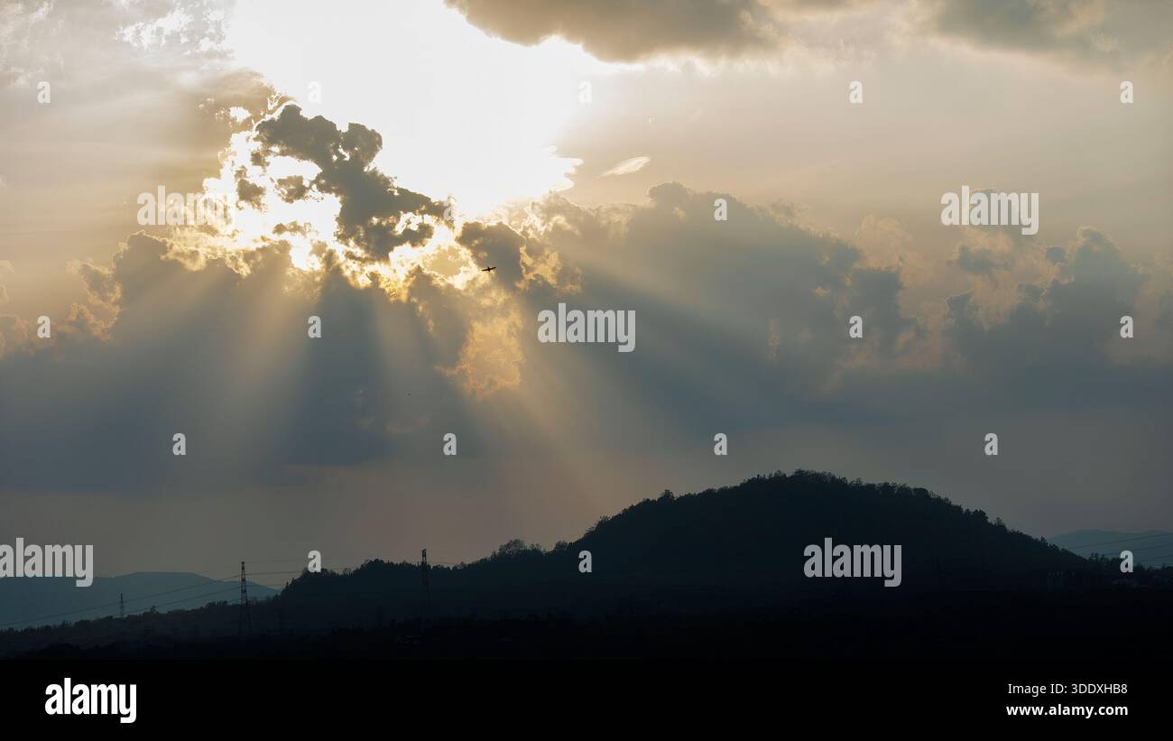 Das Flugzeug fliegt durch dramatische Sonnenstrahlen, die bei Sonnenuntergang durch dicke Wolken über einer dunklen Bergsilhouette brechen. Starker Kontrast zwischen hellem Himmel Stockfoto