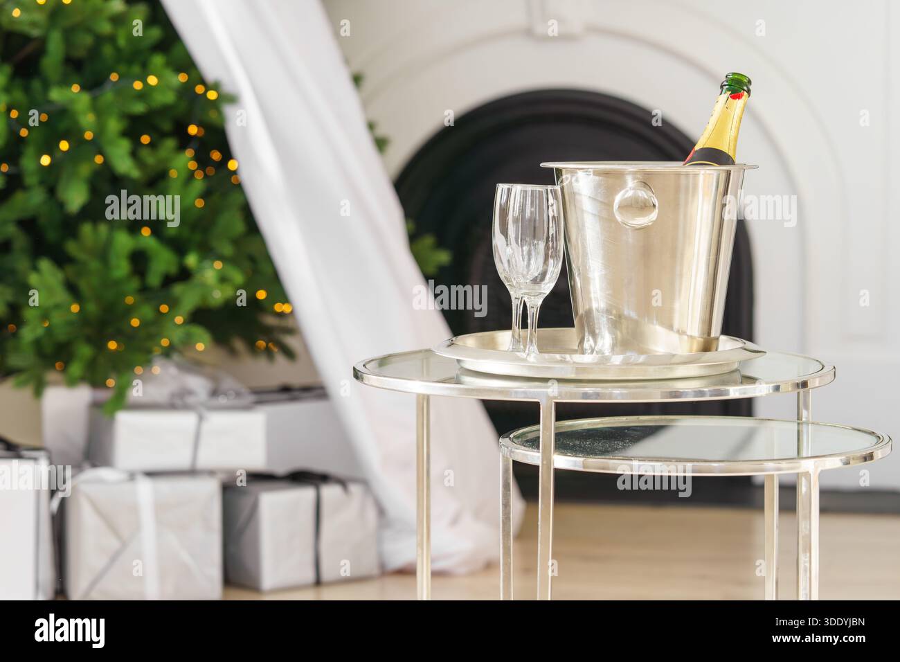 Champagnerflasche im Eiskübel aus Metall mit zwei leeren Gläsern auf Glastisch auf leuchtenden Weihnachtsbaumlichtern und Geschenken, gegenwärtiger Hintergrund. Feierlicher Sieg Stockfoto