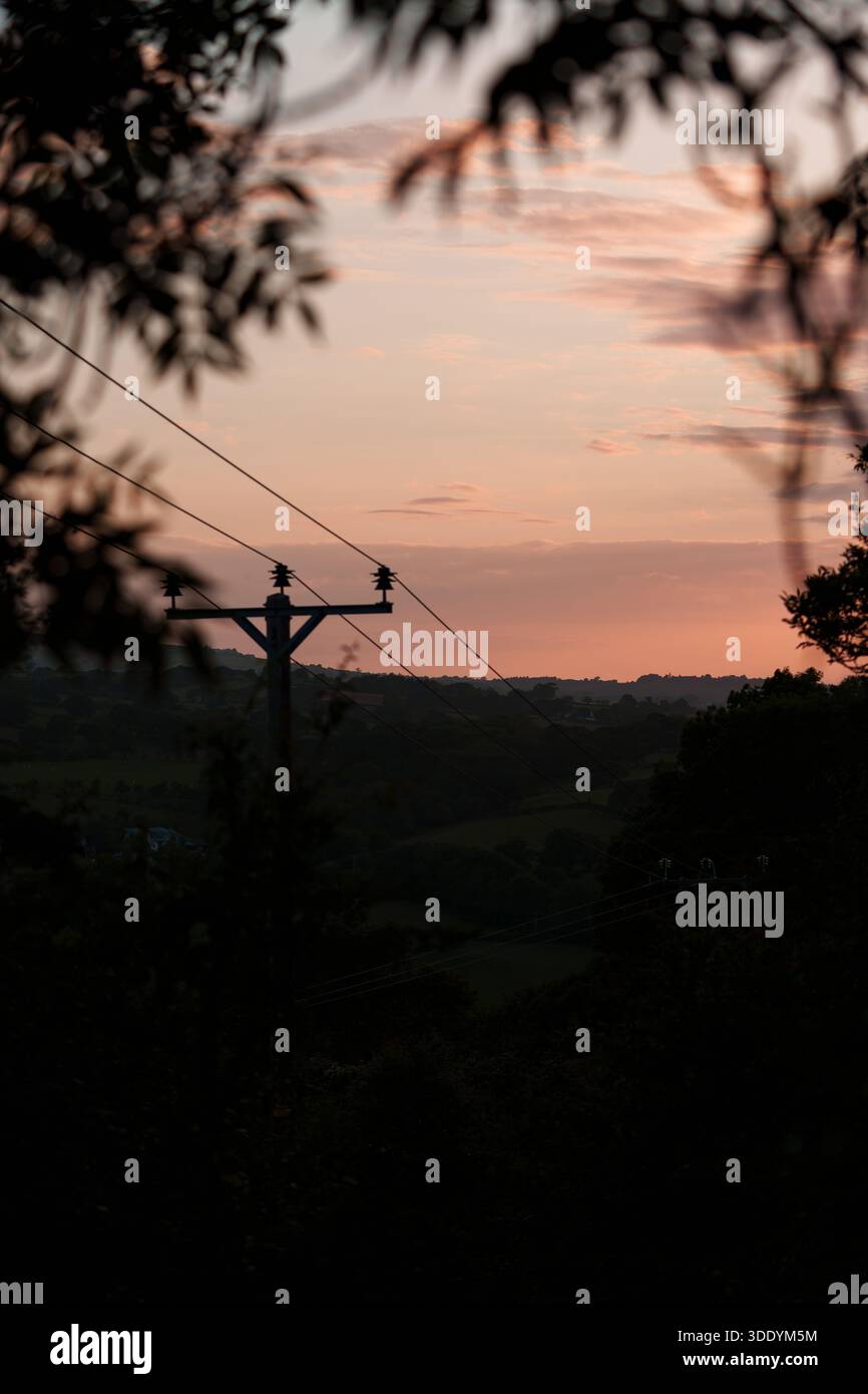 Ländliche Landschaft in Wales mit Stromleitungen gegen einen warmen Sonnenuntergang. Goldenes Licht hebt sanfte Hügel und ländliche Konturen hervor. Stockfoto