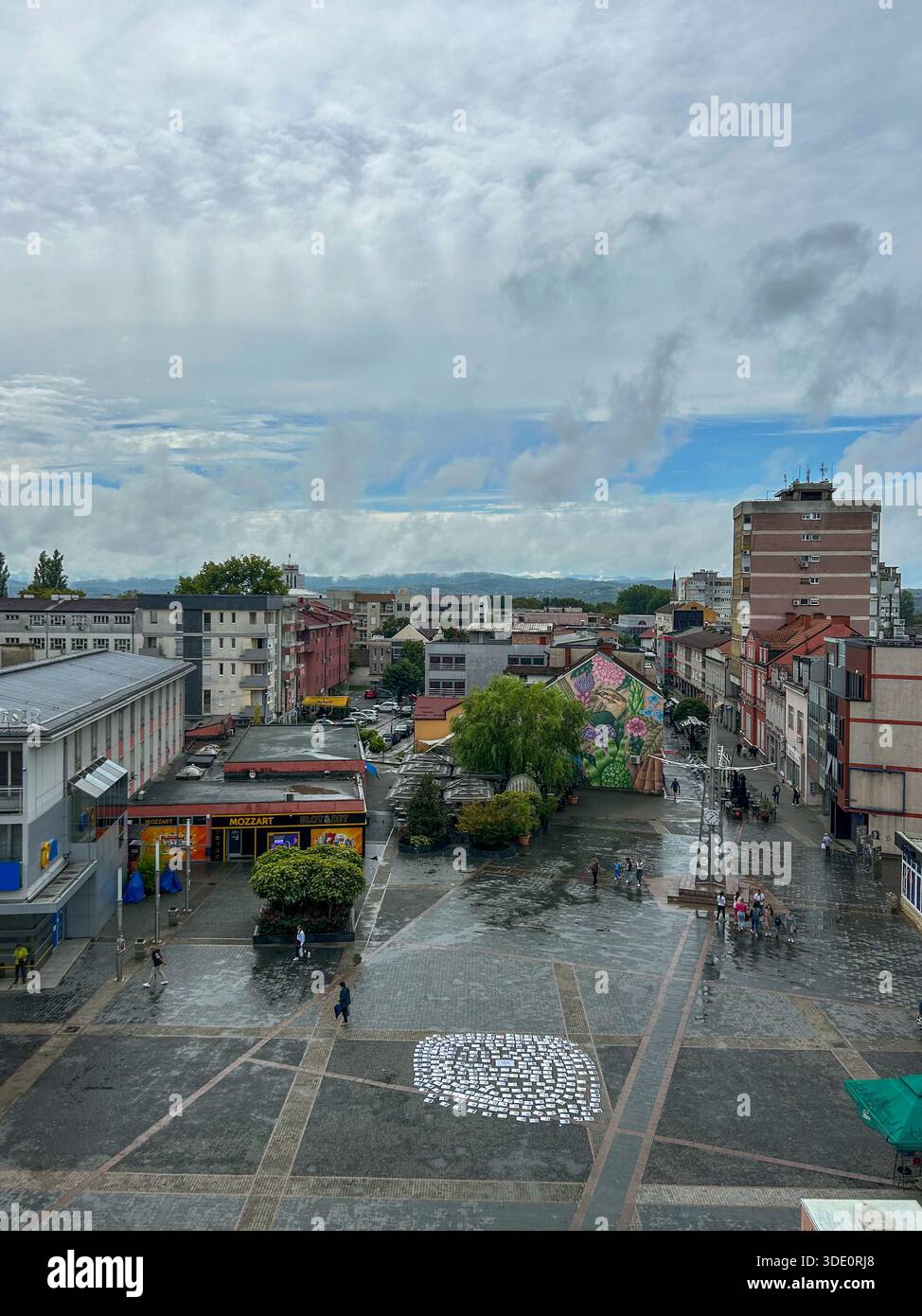 Blick auf den Stadtplatz von Prijedor nach Regen, mit städtischen Gebäuden, nassen Reflexen auf dem Bürgersteig, Fußgängern und dramatischen Wolken in der Republika Srpska - Smartphone-aufgenommenes Stockfoto