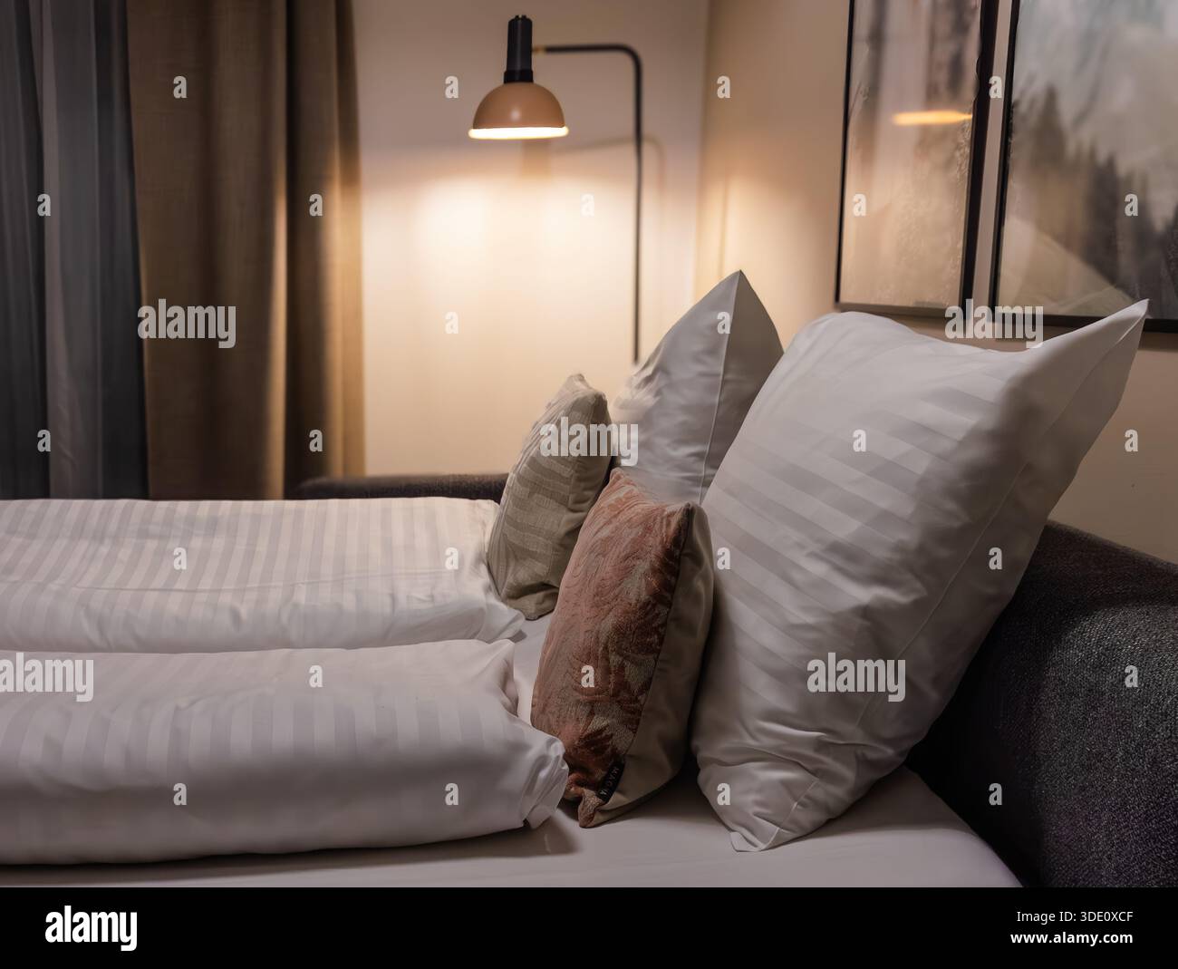 Horizontale Weitwinkelaufnahme auf Augenhöhe mit Fokus auf einem komfortablen Bett mit gestreifter weißer Bettwäsche, dekorativen Kissen und einer leuchtenden Nachttischlampe Stockfoto