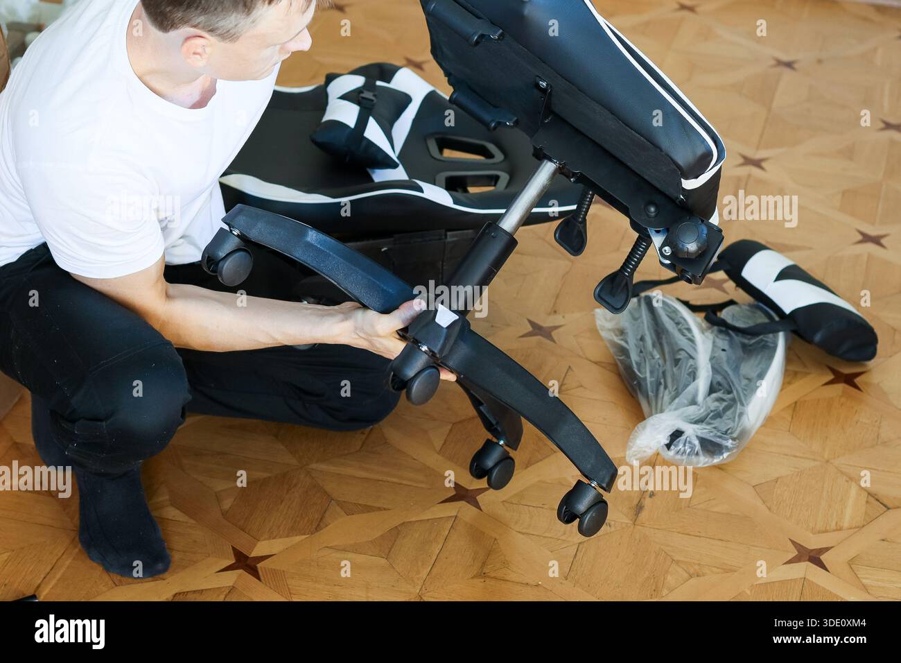 Der Squatting man befestigt schwarze Räder am Metallgestell eines Spielstuhls mit Kissen und Plastikfolie auf Holzboden. Stockfoto