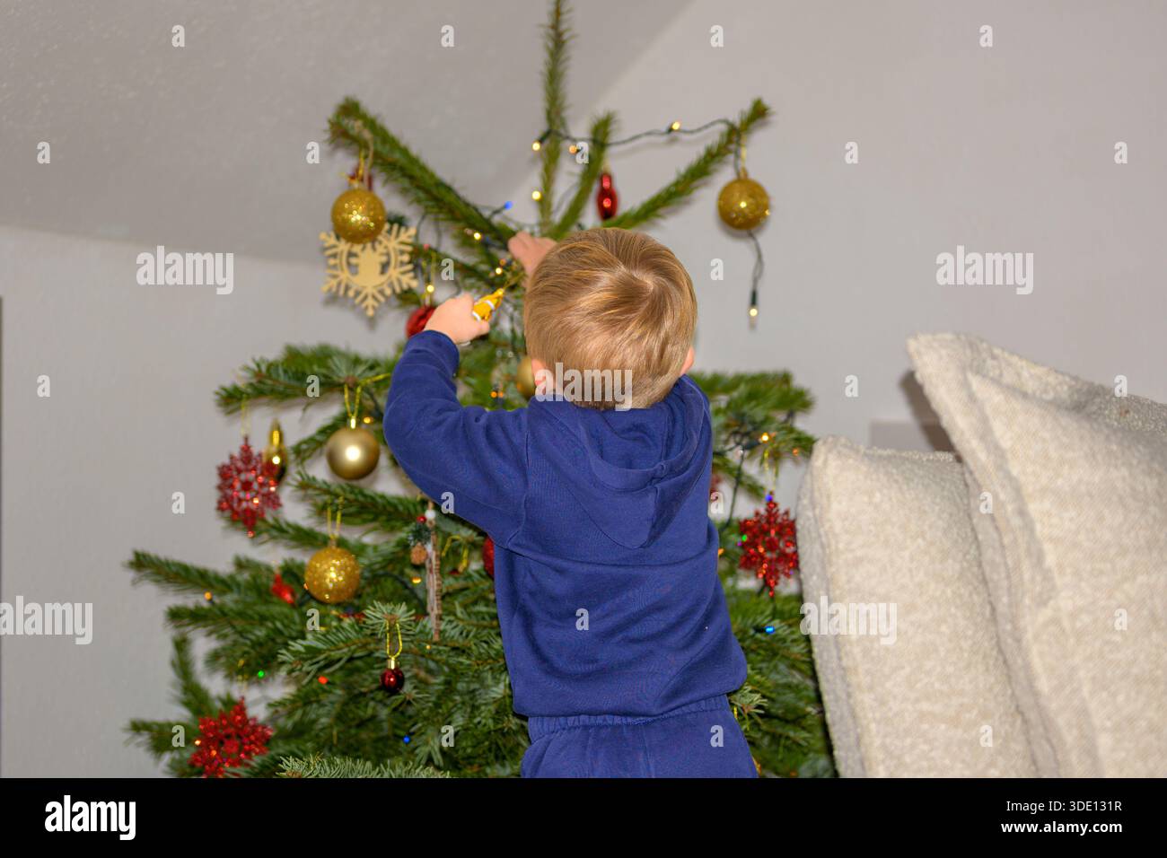 Ein Kleinkind steht mit Rückansicht und ziert einen Weihnachtsbaum mit Ornamenten und Lichtern in einer gemütlichen, festlichen Atmosphäre. Stockfoto