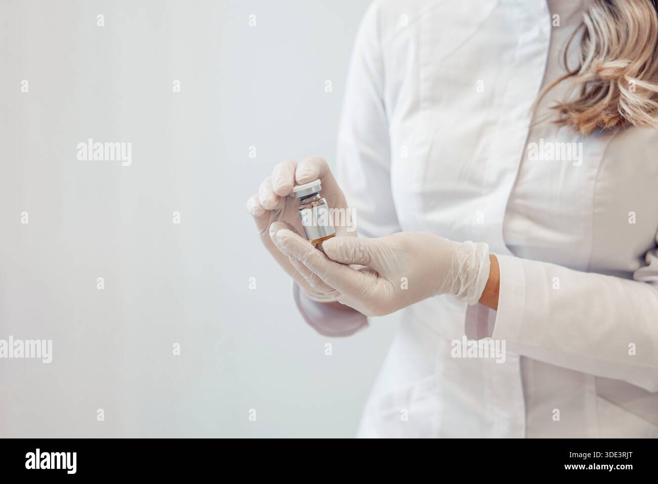 Eine Kosmetikerin in einem weißen Mantel hält eine Durchstechflasche mit Serum. Sie trägt Handschuhe und hat langes, welliges Haar. Die Einstellung ist sauber und steril und konzentriert sich auf s Stockfoto