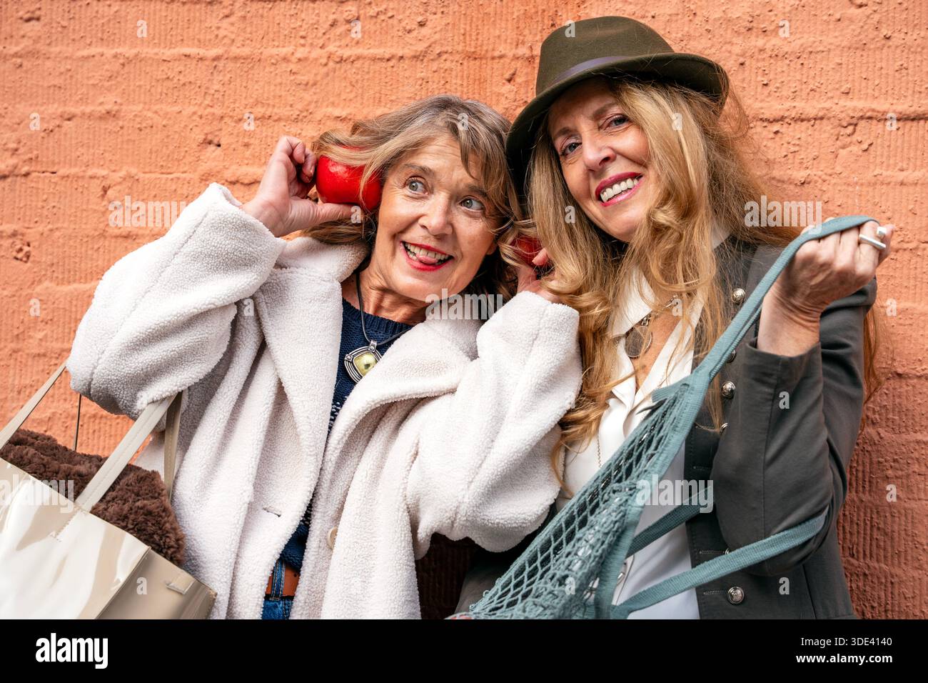 Ein helles Lächeln erleuchtet die Szene, während zwei Frauen vor einer lebendigen Ziegelwand stehen und ihren verspielten Geist mit lustigen Accessoires und Shopping-Ba zum Ausdruck bringen Stockfoto