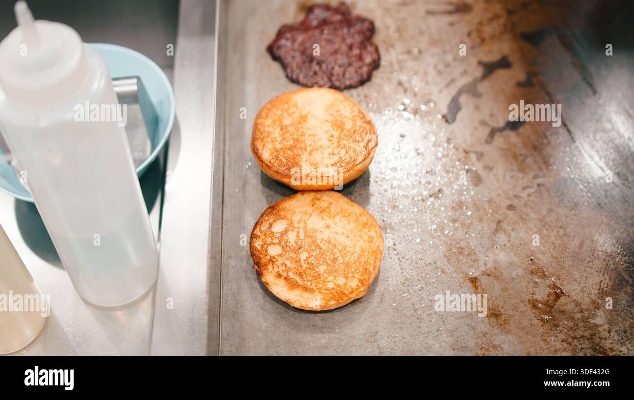 Burgerbrötchen, die zusammen mit einem Fleischpatty auf einem gewerblichen Grilltisch mit flacher Oberseite toasten Stockfoto