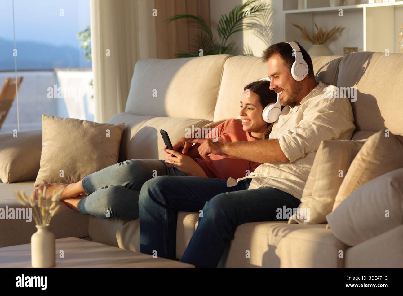 Glückliches Paar, das Kopfhörer trägt und auf einem Sofa sitzt, das zu Hause auf dem Telefon lauscht Stockfoto