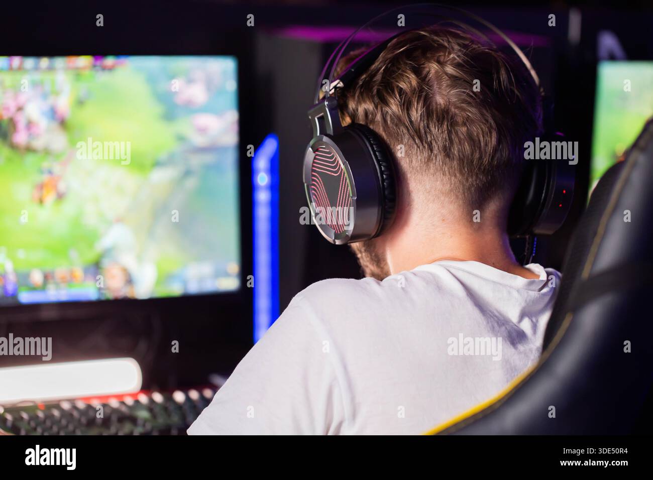 Rückansicht einer Person, junger Mann mit Kopfhörern, der an Online-Gaming-Sitzungen teilnimmt, helle Spielgrafiken auf dem Computerbildschirm im dunklen Raum, Mod Stockfoto