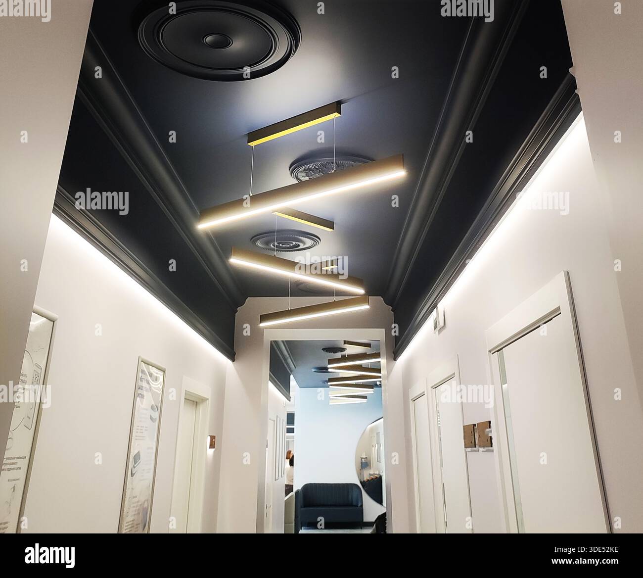 Moderne Inneneinrichtung des langen Flurs. Die dunkle Decke ist mit dekorativen Zierleisten ausgestattet. Lineare Pendelleuchten beleuchten den Raum und schaffen eine helle Atmosphäre. Anzeigen z. B. Stockfoto